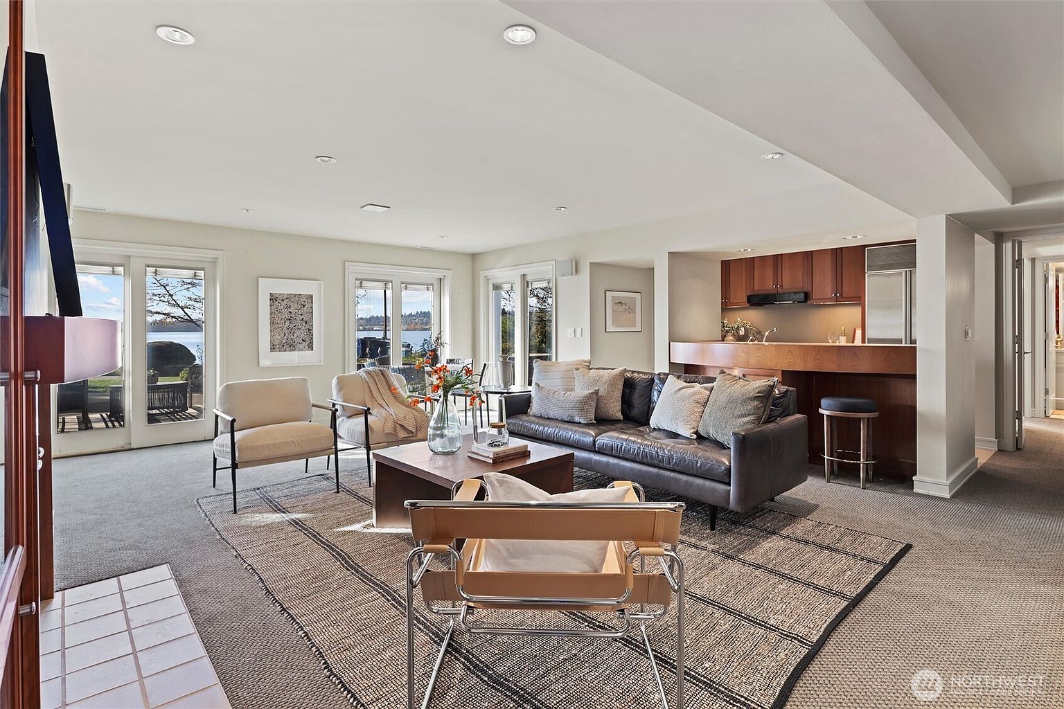 This spacious, open-concept living room features a sophisticated blend of modern and comfortable furnishings, including a dark leather sofa and a stylish leather-strapped accent chair. Large glass doors provide an expansive view of the waterfront, flooding the room with natural light and creating a seamless connection to the outdoors. The space is anchored by a textured area rug and a central wooden coffee table, complemented by a convenient wet bar area in the background.