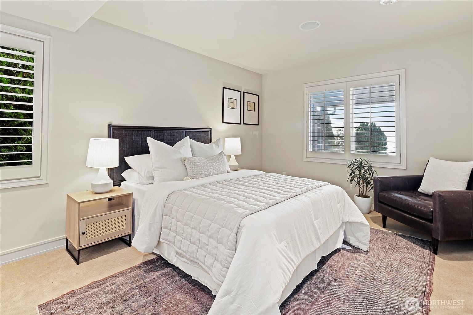 This bright and inviting bedroom features a queen-sized bed with a textured white quilt and a dark, woven headboard. The room is accented by a modern light-wood nightstand, a dark leather armchair in the corner, and a large, patterned area rug that adds warmth to the space. Natural light streams through plantation shutters on two windows, creating a serene and comfortable atmosphere.
