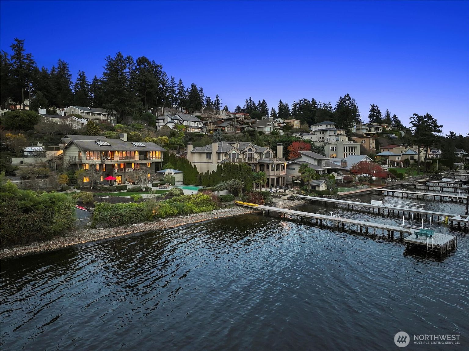This stunning aerial view captures a collection of luxury waterfront homes nestled along a lush, tree-lined hillside at dusk. The scene features multi-story residences with expansive decks, private docks extending into the calm water, and beautifully landscaped grounds, all illuminated by warm interior lighting against a deep blue twilight sky. The perspective emphasizes the prime location and the seamless integration of high-end architecture with the natural lakeside environment.