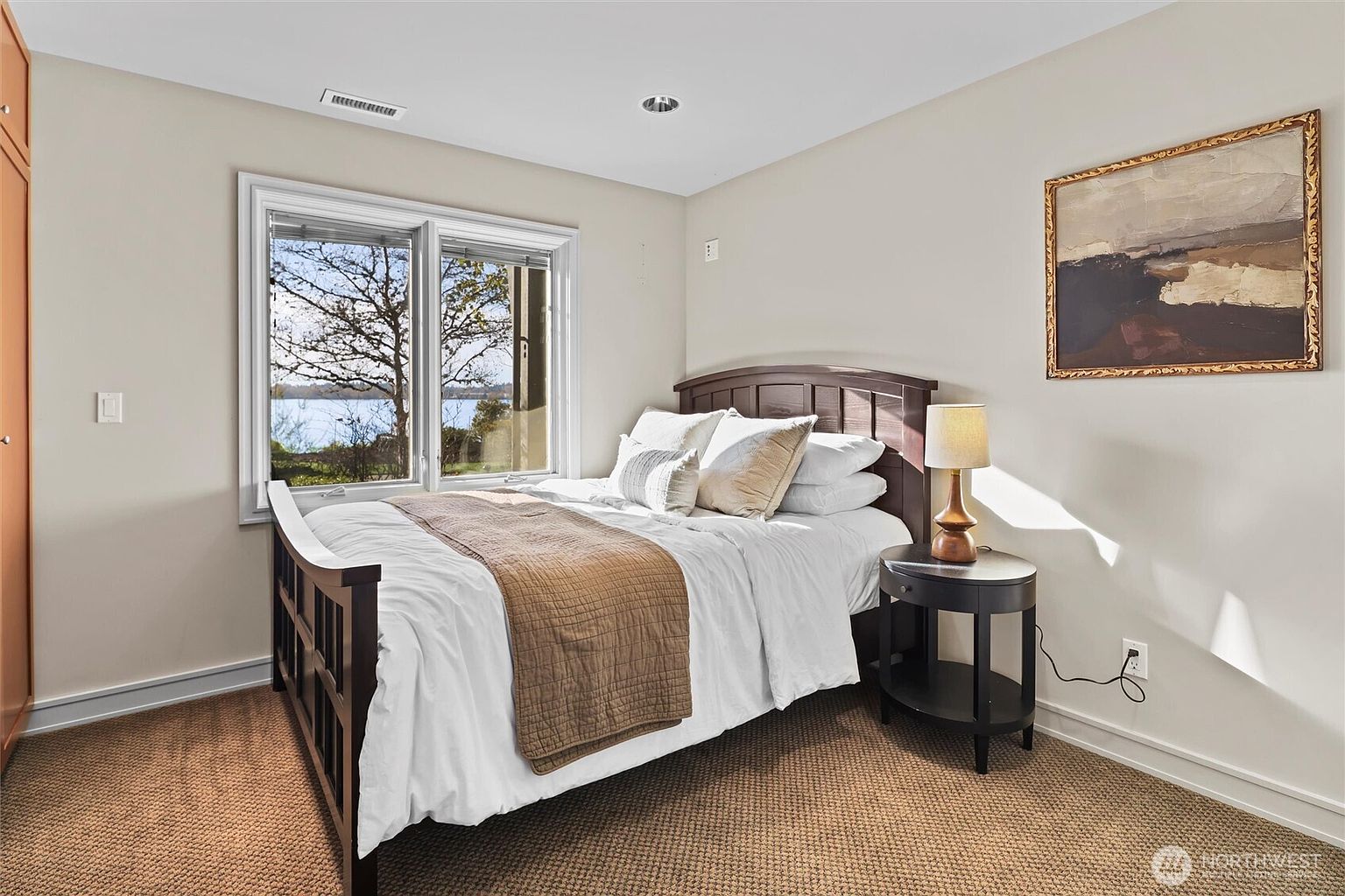 This cozy bedroom features a dark wood bed frame dressed in white linens with a brown throw, positioned next to a matching round nightstand with a warm-toned lamp. A large window provides a scenic view of the water outside, while a framed abstract painting adds a touch of artistic flair to the neutral-toned walls. The room is carpeted and maintains a calm, inviting atmosphere suitable for a guest space.