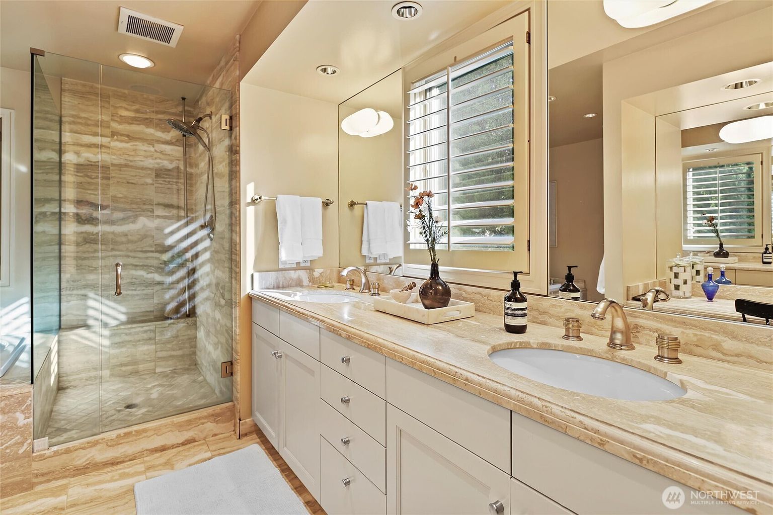 This elegant primary bathroom features a double vanity with a light-toned stone countertop and matching backsplash, complemented by white cabinetry and gold-toned fixtures. To the left, a spacious walk-in shower is enclosed by glass and finished with warm-toned, veined stone tiles that create a luxurious, spa-like atmosphere. The room is brightened by large mirrors and recessed lighting, offering a clean and sophisticated aesthetic.