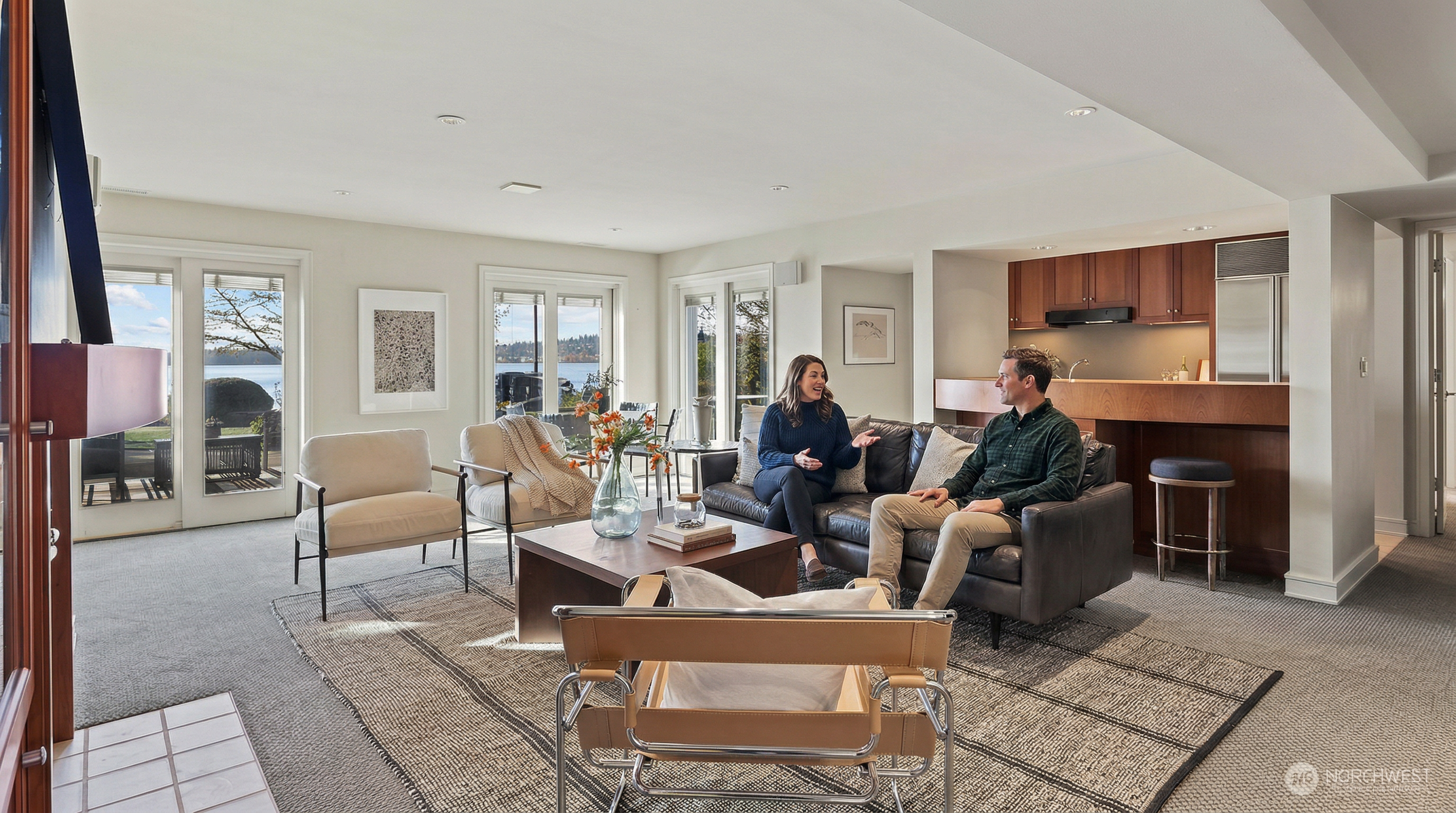 This spacious, open-concept living room features a sophisticated blend of modern and comfortable furnishings, including a dark leather sofa and a stylish leather-strapped accent chair. Large glass doors provide an expansive view of the waterfront, flooding the room with natural light and creating a seamless connection to the outdoors. The space is anchored by a textured area rug and a central wooden coffee table, complemented by a convenient wet bar area in the background.