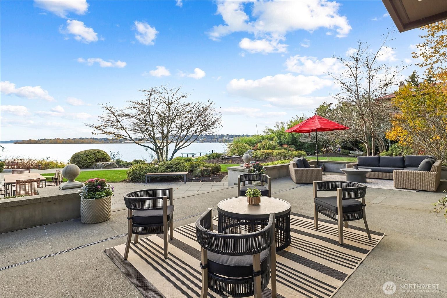 This expansive stone patio offers a luxurious outdoor living space with stunning, unobstructed views of the lake and distant shoreline. The area is furnished with comfortable lounge seating, a dining set, and a vibrant red umbrella, all set against a backdrop of mature trees and manicured landscaping. The scene captures a serene, high-end waterfront lifestyle perfect for entertaining or relaxing.