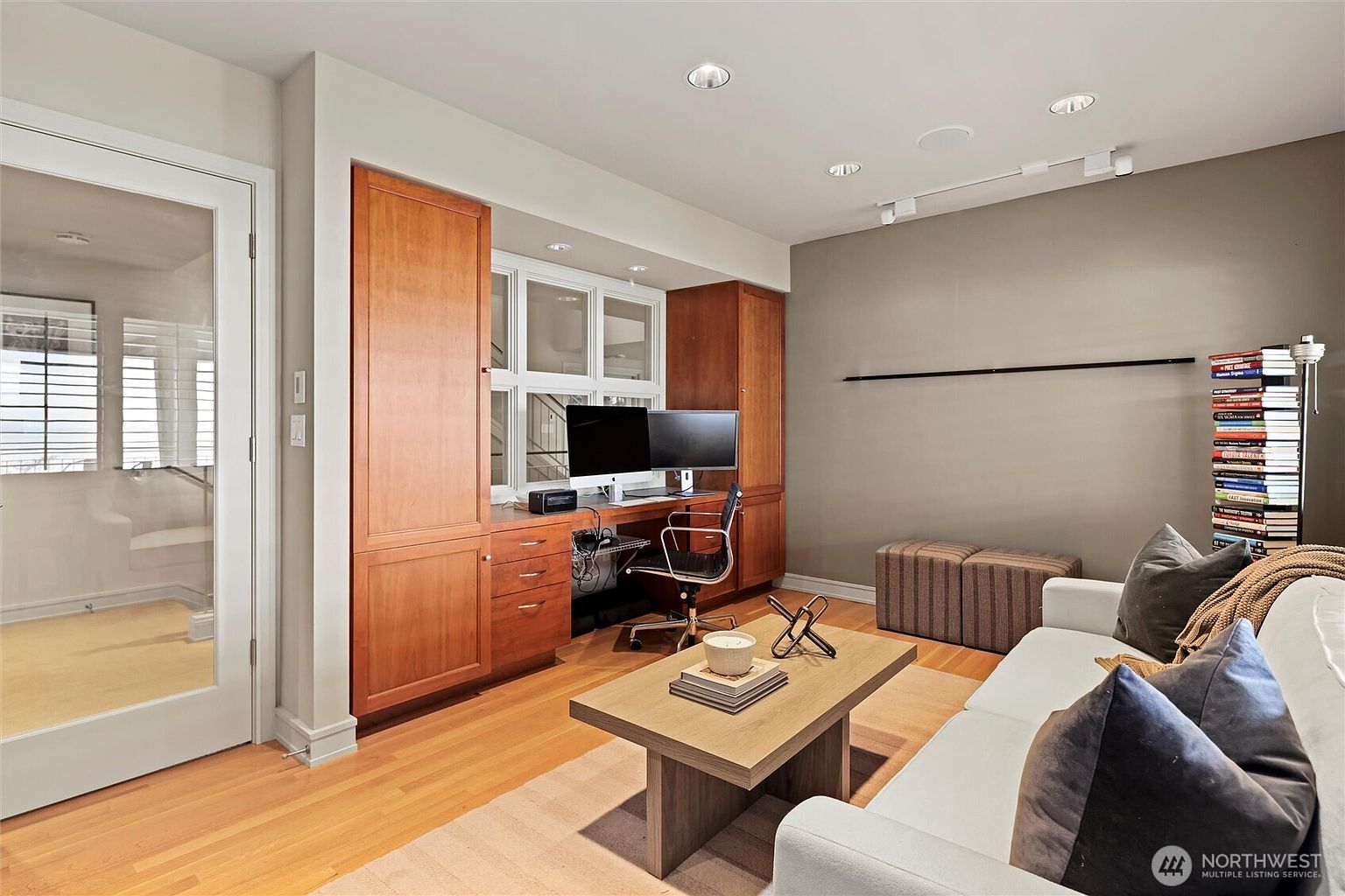 This well-organized home office features custom built-in wooden cabinetry surrounding a desk area with dual monitors, creating a functional workspace. The room includes a comfortable seating area with a sofa and coffee table, making it a versatile space for both work and relaxation. The warm wood flooring, neutral wall color, and natural light from the window create a professional yet inviting atmosphere.