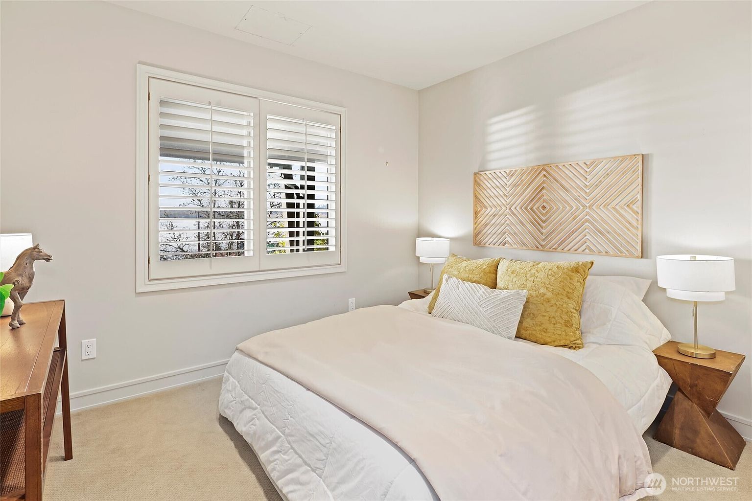 This cozy bedroom features a neutral color palette with a comfortable bed dressed in white linens and accented by textured yellow throw pillows. A decorative wooden wall art piece hangs above the headboard, while plantation shutters on the window allow for soft, natural light to filter into the room. The space is completed by two matching bedside tables with modern lamps and a small decorative horse figurine on a side console, creating a warm and inviting atmosphere.