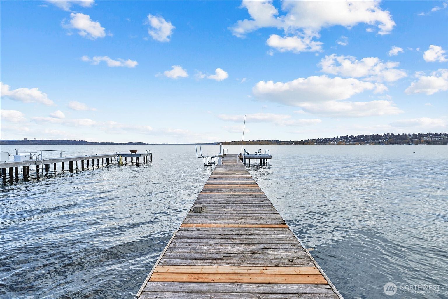 This image captures a long, weathered wooden dock extending out into a calm, expansive lake under a bright blue sky with scattered clouds. The perspective is from the shore looking out, emphasizing the peaceful waterfront setting and the direct access to the water. The scene conveys a serene and inviting atmosphere, highlighting the property's prime location for boating or relaxation.