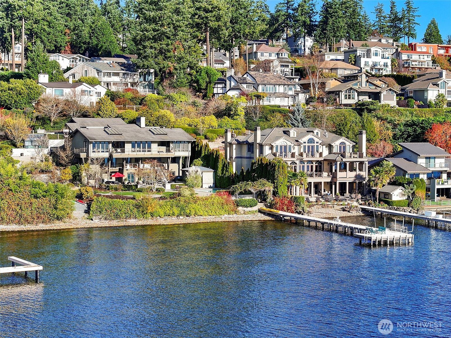 This elevated, wide-angle shot captures a stunning waterfront property featuring two large, multi-story luxury homes nestled on a lush, tree-covered hillside. The scene highlights the expansive private docks extending into the calm blue water, manicured landscaping, and a tiered architectural design that maximizes the scenic lake views. The overall impression is one of high-end, serene lakeside living in a prestigious residential neighborhood.