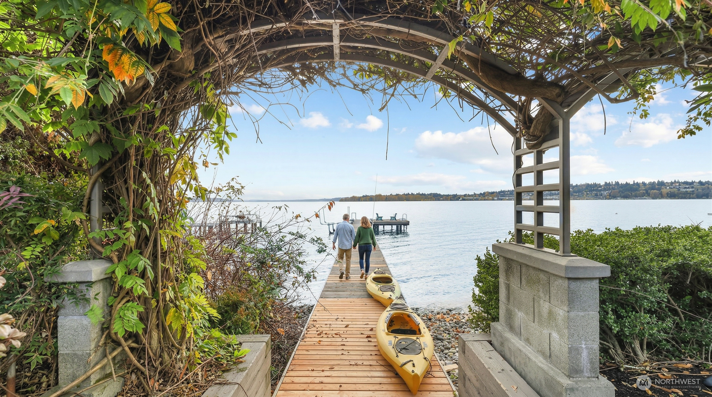 This picturesque view captures a wooden dock extending from a lush, vine-covered arbor toward a serene lake. Two yellow kayaks rest on the dock, inviting outdoor recreation, while the surrounding greenery and calm water create a tranquil, inviting atmosphere. The perspective is framed through the arbor, drawing the viewer's eye directly toward the expansive waterfront horizon.