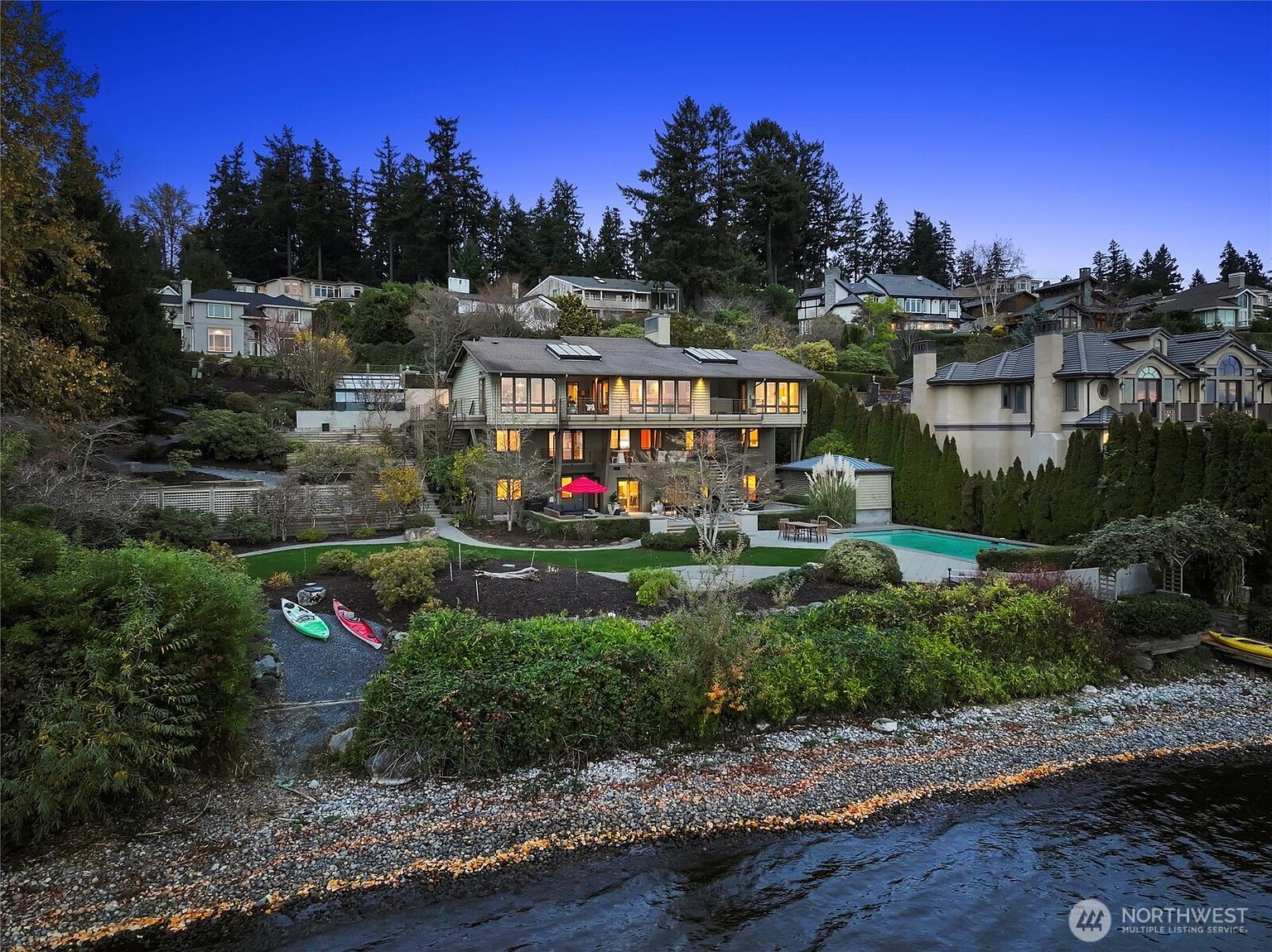 This stunning waterfront property features a multi-level residence with expansive windows and balconies overlooking a lush, tiered backyard. The outdoor space includes a pristine swimming pool, manicured lawn, and direct access to the rocky shoreline, creating a serene and luxurious atmosphere. Captured from an elevated perspective, the twilight lighting enhances the home's architectural elegance and inviting ambiance.