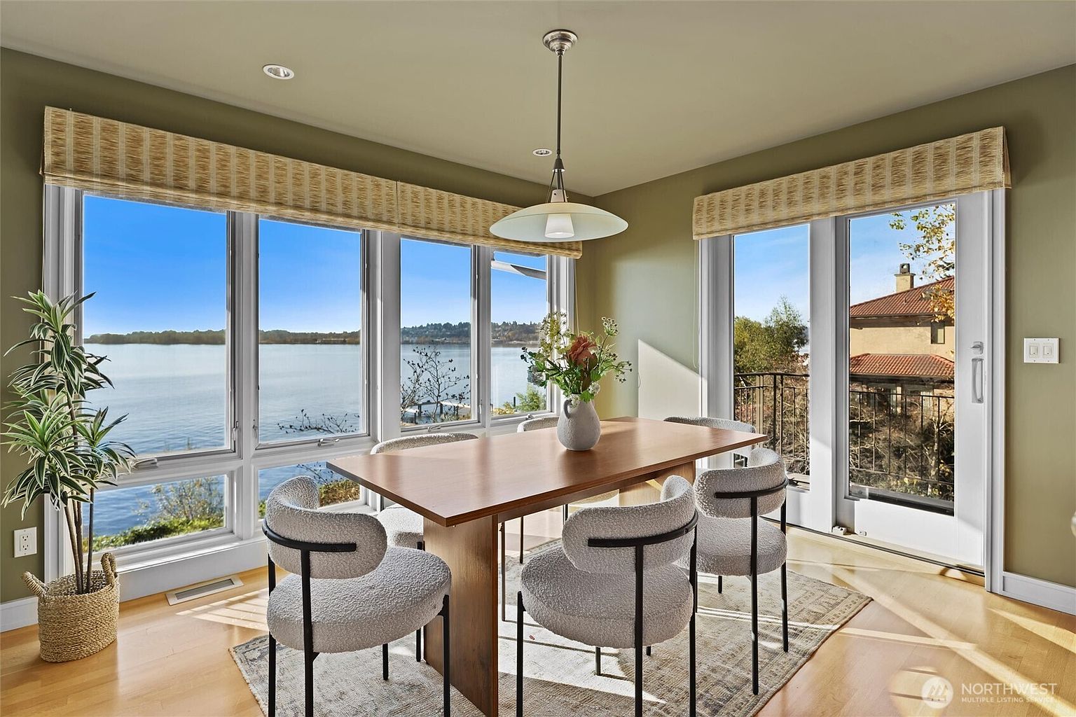 This bright and airy dining room features a modern wooden table surrounded by four textured, light-gray upholstered chairs. Large floor-to-ceiling windows and a glass door provide expansive, scenic views of the water, filling the space with natural light. The room is accented by a contemporary pendant light fixture and a patterned area rug, creating a sophisticated and inviting atmosphere.