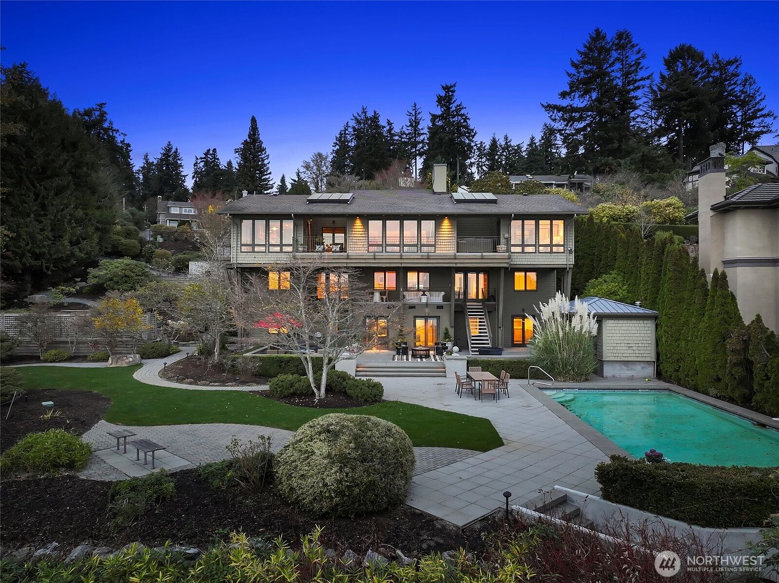 This stunning rear view of a multi-story residence showcases a sophisticated architectural design with expansive windows, multiple balconies, and a seamless transition to a beautifully landscaped backyard. The property features a large stone patio, a pristine swimming pool, and manicured lawns, all set against a backdrop of lush, mature trees under a twilight sky. The warm interior lighting creates an inviting and luxurious atmosphere, highlighting the home's potential for elegant outdoor entertaining.