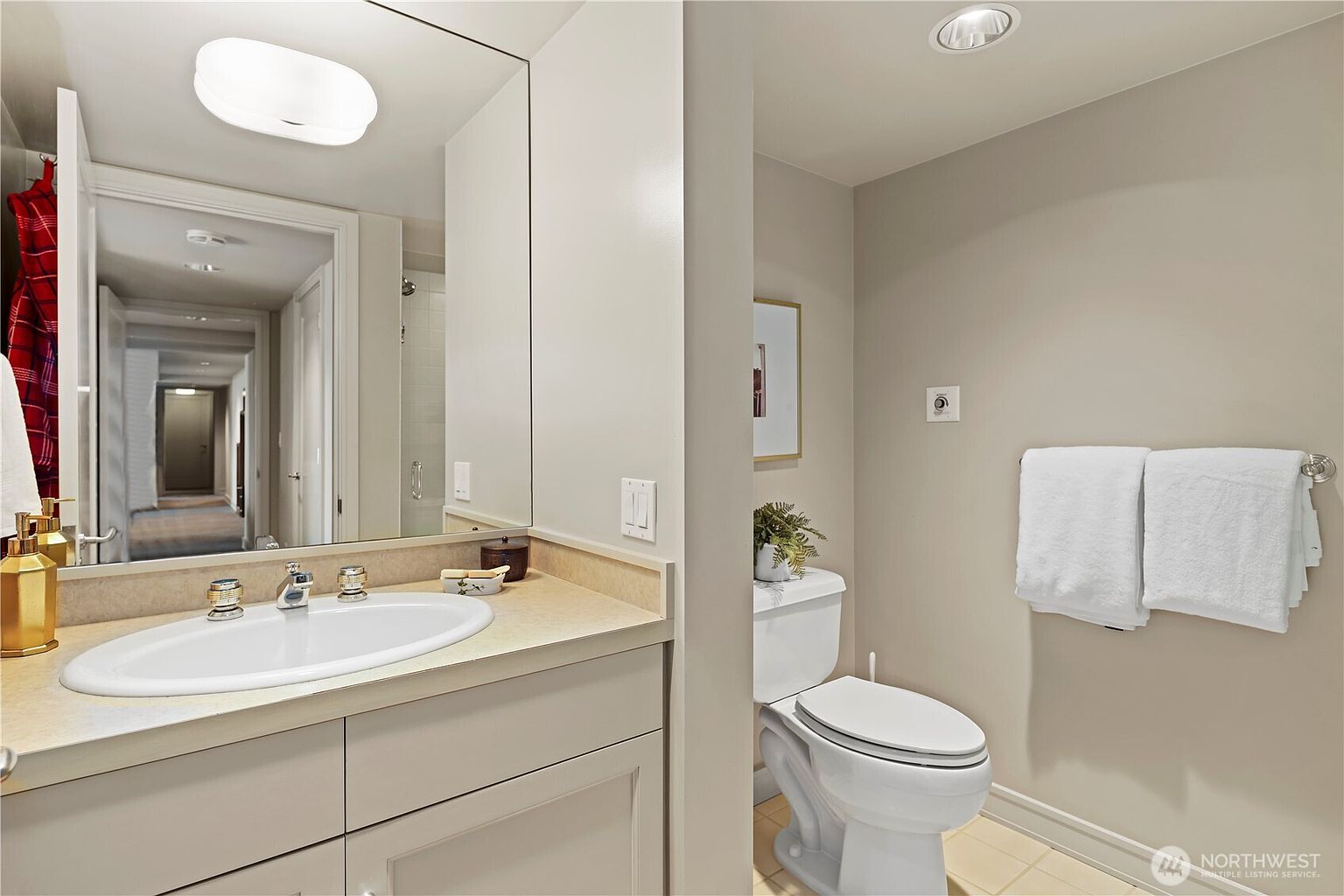 This clean and functional bathroom features a neutral color palette with a vanity topped by a light-colored countertop and a large wall-mounted mirror. To the right, a standard white toilet is positioned next to a towel rack holding two white towels, creating a simple and tidy aesthetic. The perspective is a straight-on shot that captures the vanity and toilet area, reflecting the hallway in the mirror to provide a sense of depth.