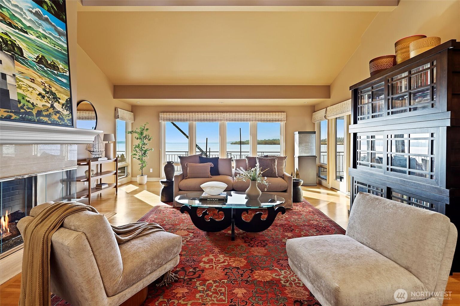 This elegant living room features a vaulted ceiling and a large, central fireplace adorned with a coastal-themed painting, creating a sophisticated yet relaxed atmosphere. A plush sofa and two armchairs are arranged around a unique glass-topped coffee table on a vibrant, patterned area rug, all facing a wall of windows that offer a stunning, unobstructed view of the water. The space is further enhanced by a large, dark wood bookshelf unit and warm, natural lighting, making it an inviting area for both relaxation and entertaining.