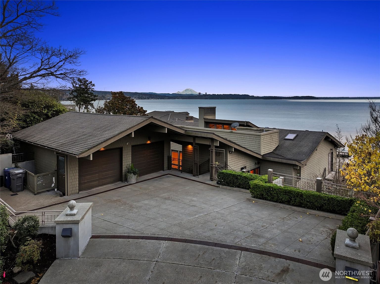 This elevated, wide-angle shot captures the front exterior of a contemporary waterfront home, showcasing a spacious concrete driveway leading to a two-car garage. The architectural design features clean lines and shingled siding, perfectly positioned to frame a breathtaking panoramic view of the bay and a distant snow-capped mountain. The scene is set during the golden hour, with a deep blue sky contrasting against the warm, inviting glow emanating from the home's interior.