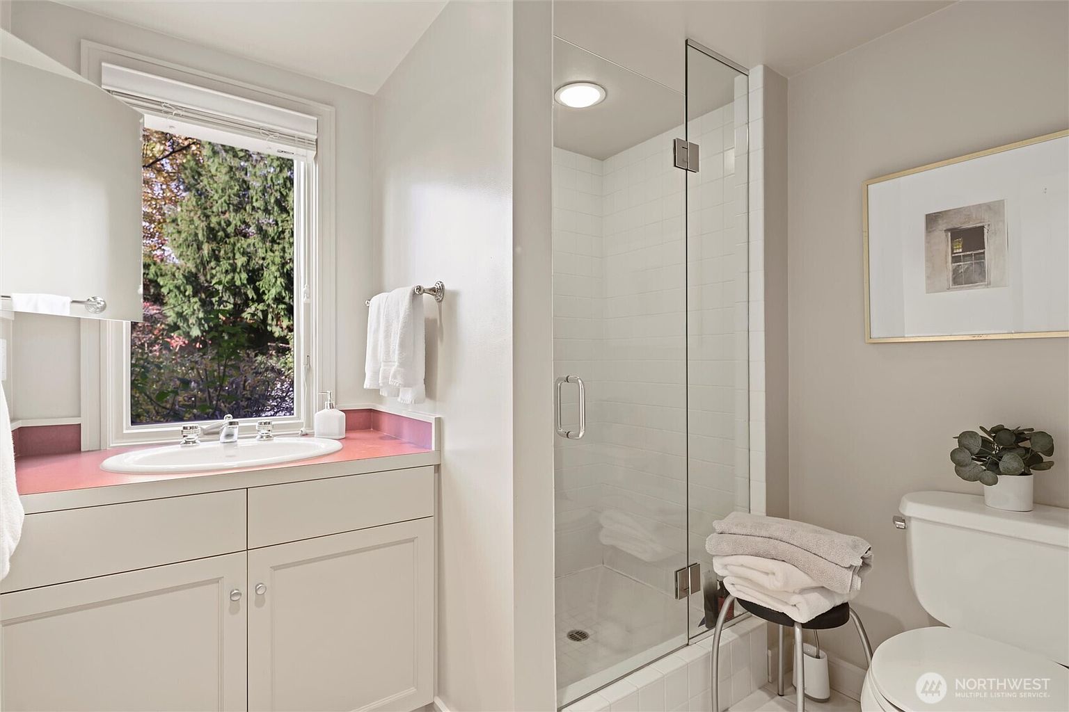 This bright and functional bathroom features a vanity with a pink countertop, a glass-enclosed walk-in shower with white subway tiles, and a toilet. A window above the sink provides natural light and a view of the outdoors, while a framed piece of art adds a decorative touch to the neutral-toned walls. The space is clean and well-maintained, offering a practical layout for a guest or secondary bathroom.