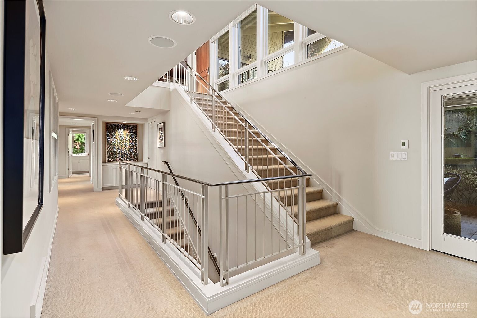 This bright and airy interior space features a prominent staircase with a modern metal railing and plush carpeting. The hallway leads to a secondary room in the distance, while large windows above the stairs allow for ample natural light to fill the area. The neutral color palette and clean lines create a sophisticated and welcoming atmosphere.