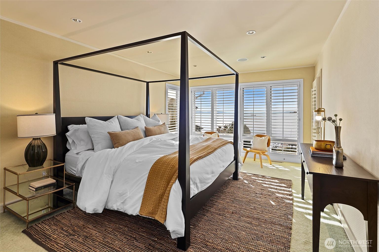 This elegant primary bedroom features a striking black four-poster canopy bed as the central focal point, dressed in crisp white linens and a warm mustard-colored throw. The room is bathed in natural light from large windows with white plantation shutters that offer a serene view of the water. A glass-topped nightstand and a dark wood console table add sophisticated contrast to the soft, neutral-toned walls and plush carpeting.
