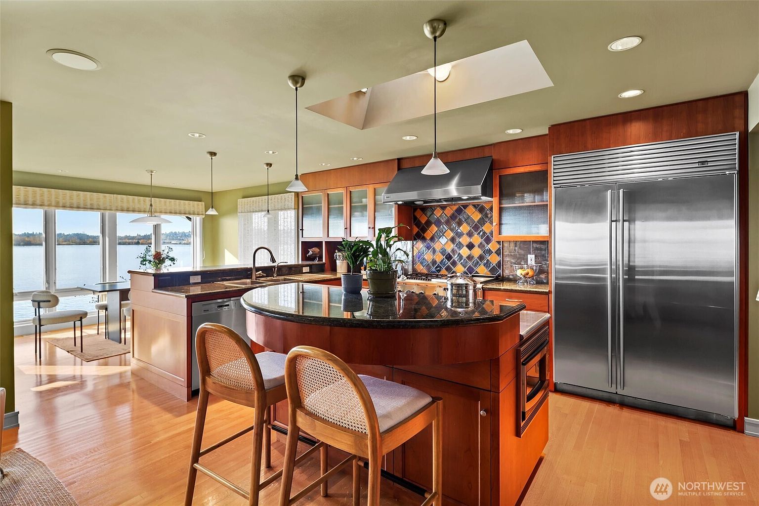 This spacious, warm-toned kitchen features rich wood cabinetry, a curved granite-topped island with bar seating, and high-end stainless steel appliances, including a large side-by-side refrigerator. A striking mosaic tile backsplash adds a pop of color, while a skylight and large windows overlooking the water provide abundant natural light. The open-concept design creates an inviting atmosphere, blending functional culinary space with scenic views.