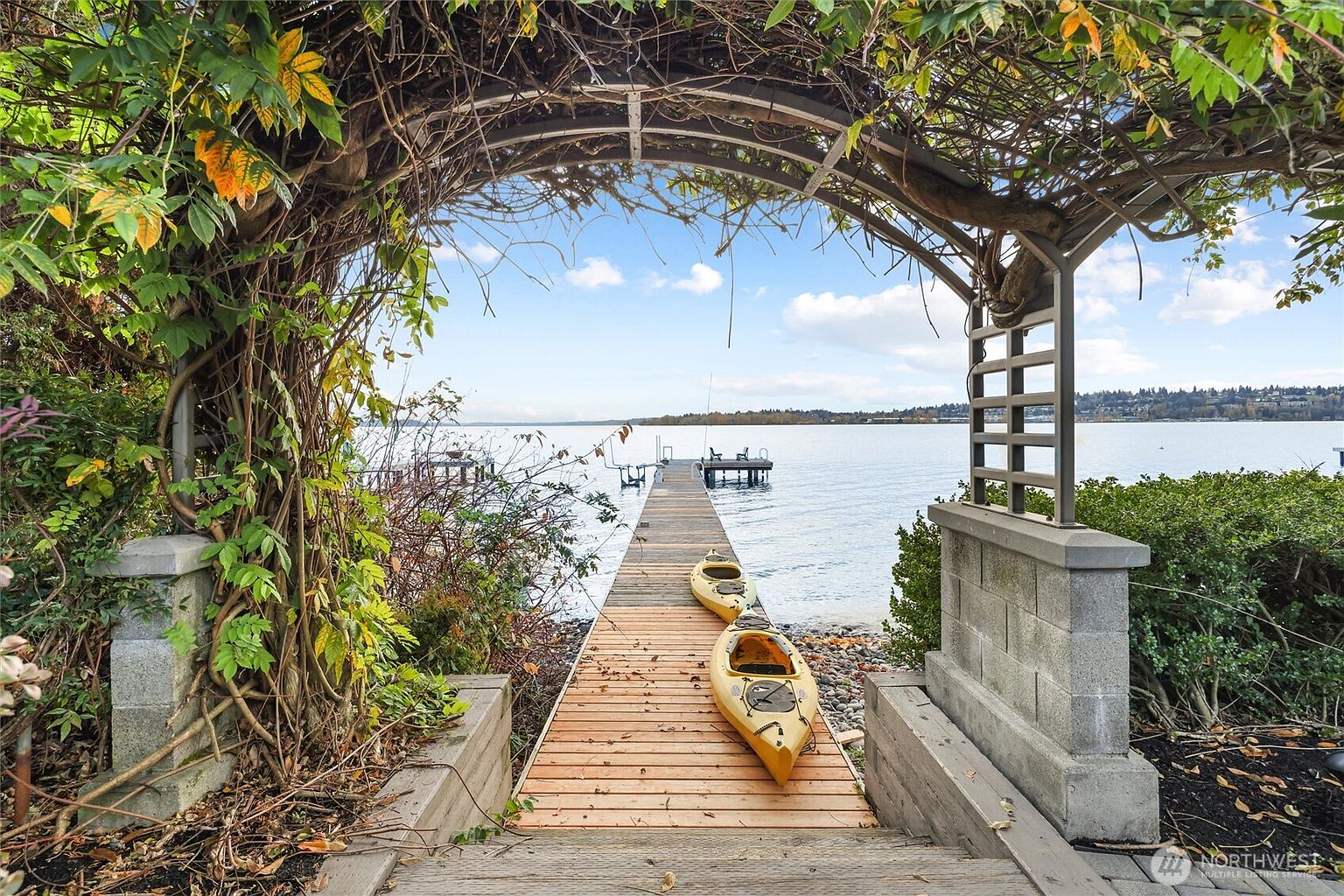 This picturesque view captures a wooden dock extending from a lush, vine-covered arbor toward a serene lake. Two yellow kayaks rest on the dock, inviting outdoor recreation, while the surrounding greenery and calm water create a tranquil, inviting atmosphere. The perspective is framed through the arbor, drawing the viewer's eye directly toward the expansive waterfront horizon.