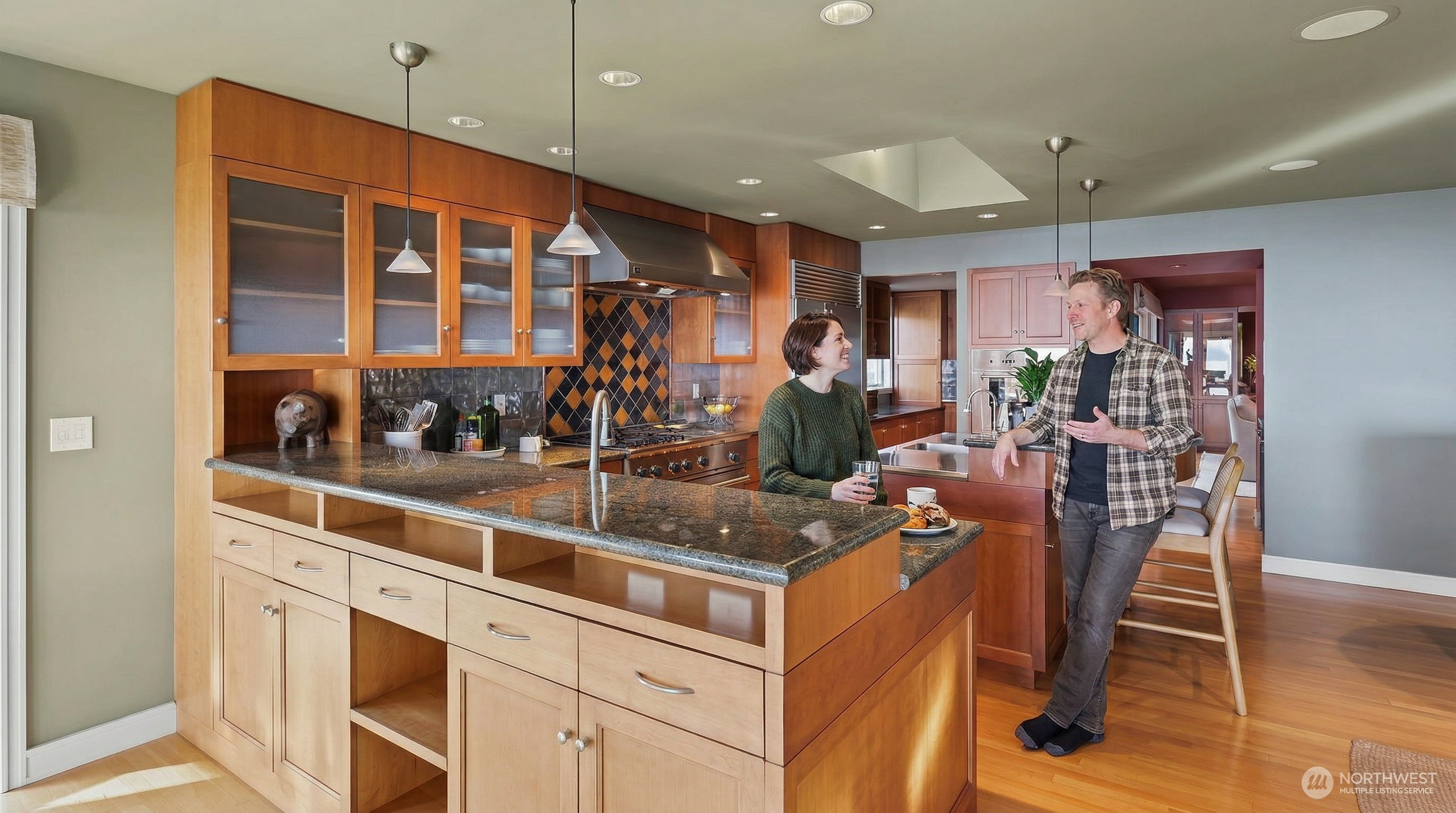 This spacious, warm-toned kitchen features natural wood cabinetry, dark granite countertops, and a large central island with integrated bar seating. The space is well-lit by recessed lighting and stylish pendant fixtures, with a unique tiled backsplash adding a touch of character. The open-concept layout flows seamlessly into the adjacent living area, creating an inviting and functional environment for cooking and entertaining.