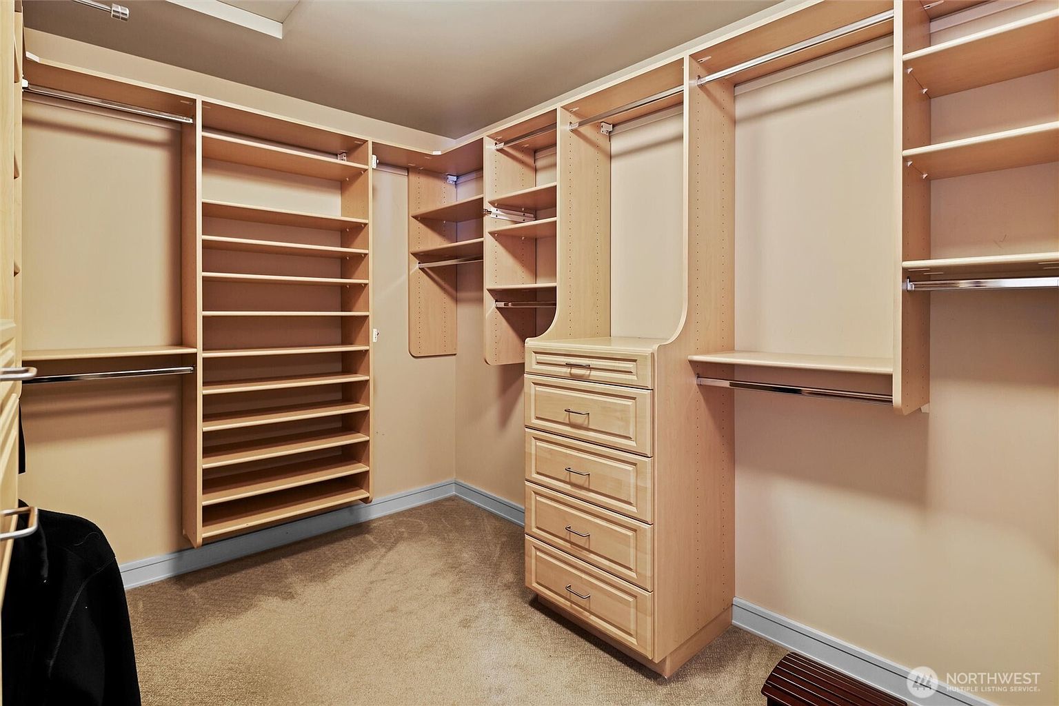This spacious walk-in closet features custom light-wood shelving and cabinetry, providing ample storage for clothing and accessories. The layout includes a central bank of drawers, multiple hanging rods, and open shelving units, all set against neutral-toned walls and carpeted flooring. The perspective is a wide-angle shot from the entrance, showcasing the organized and functional design of the storage space.