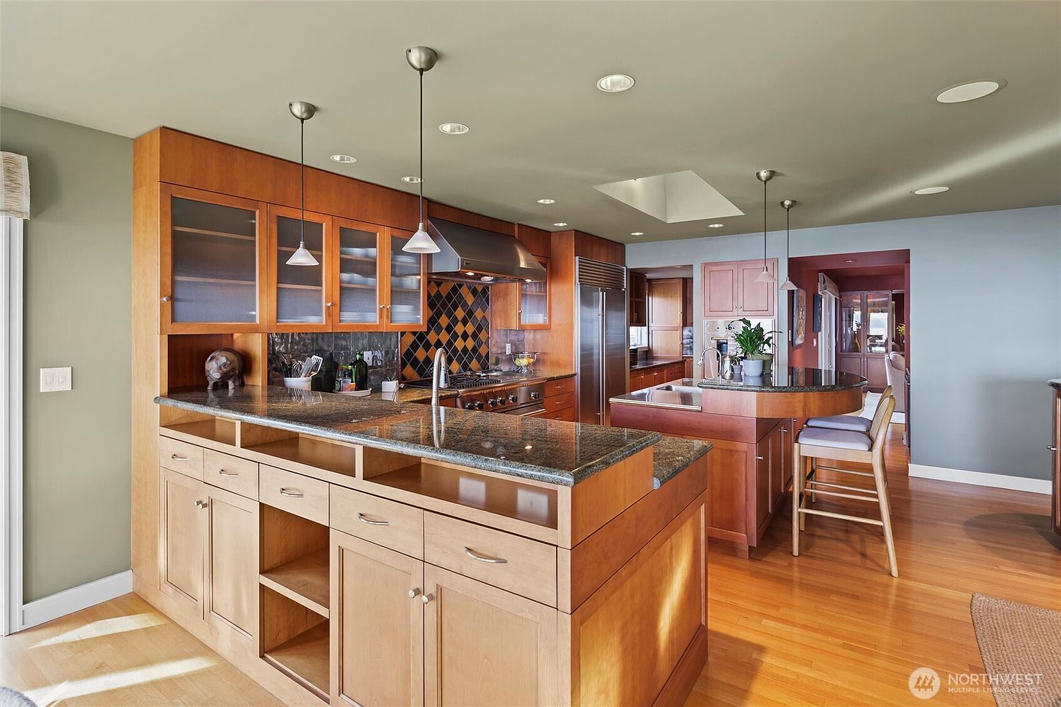 This spacious, warm-toned kitchen features natural wood cabinetry, dark granite countertops, and a large central island with integrated bar seating. The space is well-lit by recessed lighting and stylish pendant fixtures, with a unique tiled backsplash adding a touch of character. The open-concept layout flows seamlessly into the adjacent living area, creating an inviting and functional environment for cooking and entertaining.