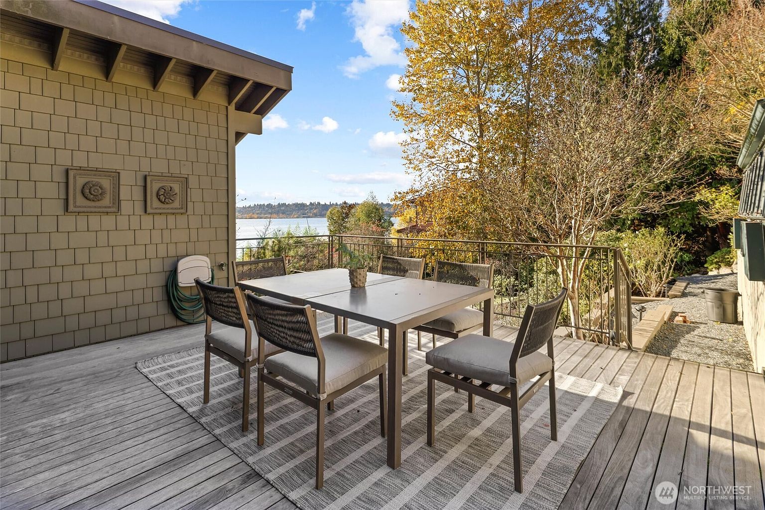 This inviting outdoor deck features a modern dining set placed atop a striped area rug, perfect for al fresco meals while overlooking a scenic waterfront view. The space is framed by a wooden shingle-sided house wall adorned with decorative circular plaques and a metal railing that provides safety without obstructing the natural landscape. The perspective captures a serene, elevated vantage point that highlights the seamless transition between indoor comfort and the beauty of the surrounding trees and water.