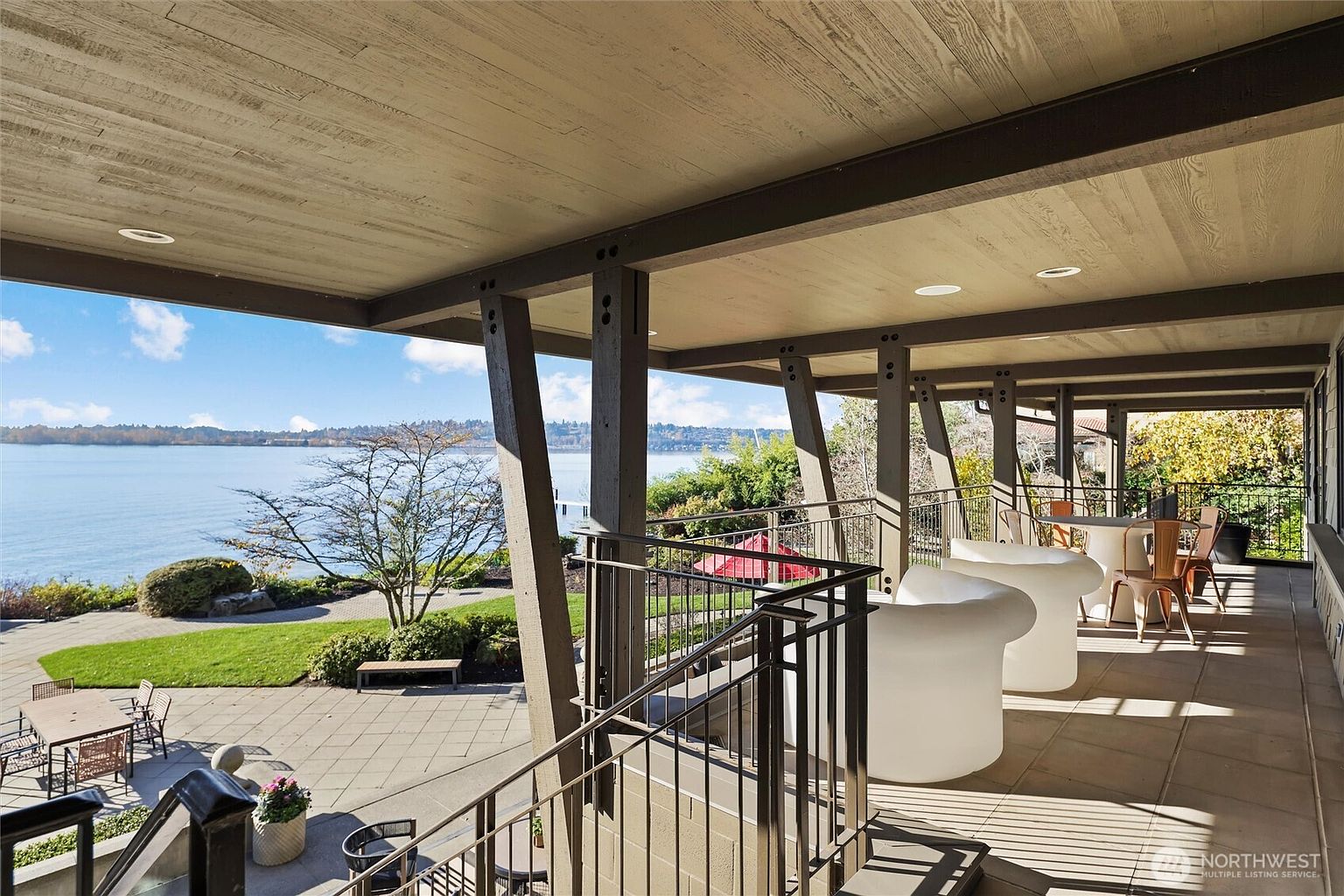 This expansive, covered deck offers a stunning, elevated view of a serene lake and lush, landscaped grounds. The space features modern, sculptural white lounge chairs and a dining area, all set against a backdrop of architectural support beams and a sleek metal railing. The perspective captures the seamless transition between the sophisticated outdoor living area and the natural beauty of the waterfront setting.