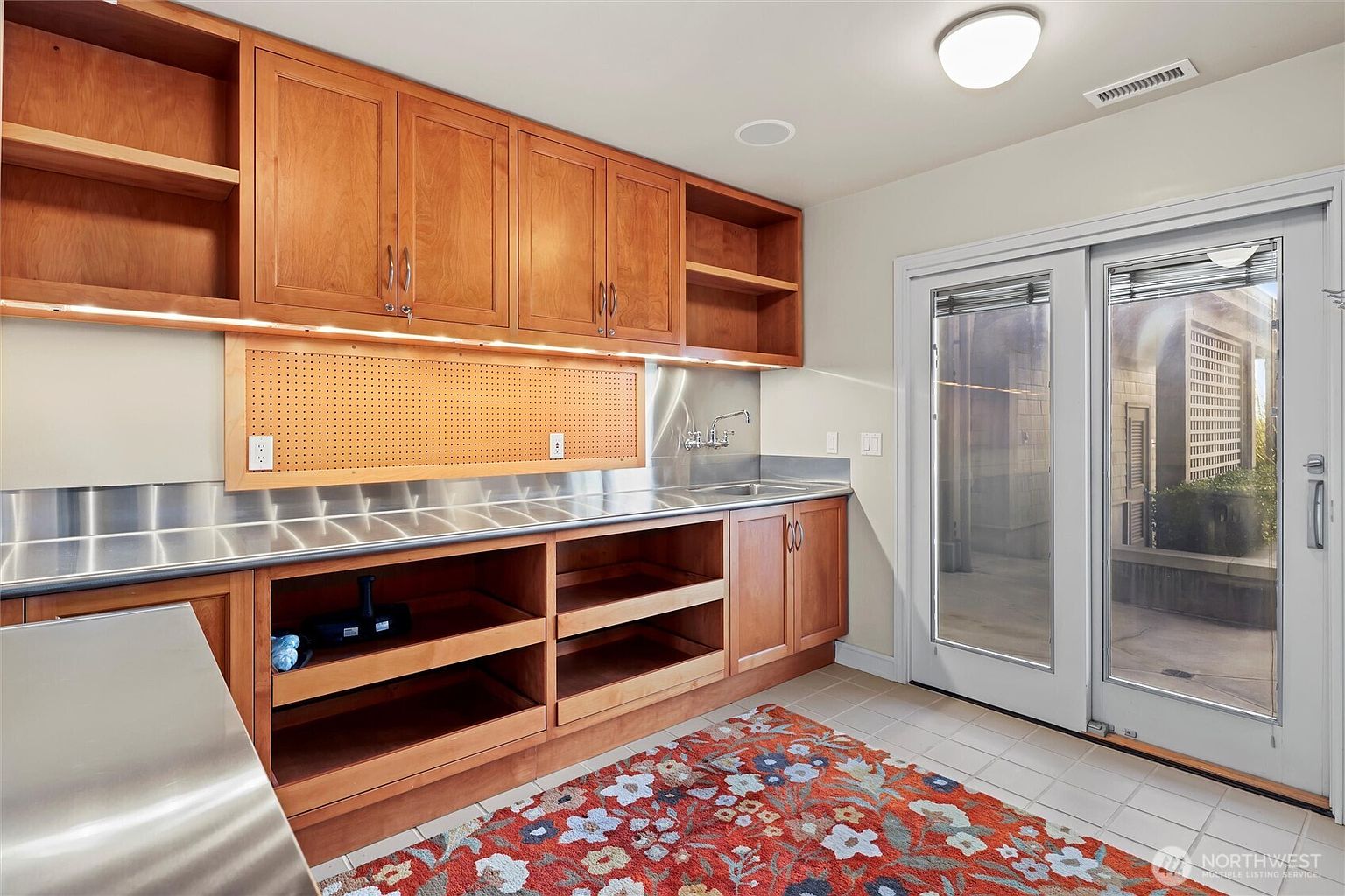 This functional laundry or utility room features warm-toned wooden cabinetry, a durable stainless steel countertop, and a built-in sink. A pegboard backsplash adds a practical touch, while the glass-paned double doors provide natural light and direct access to an outdoor patio area. The space is finished with a colorful patterned rug, creating a blend of industrial utility and homey comfort.