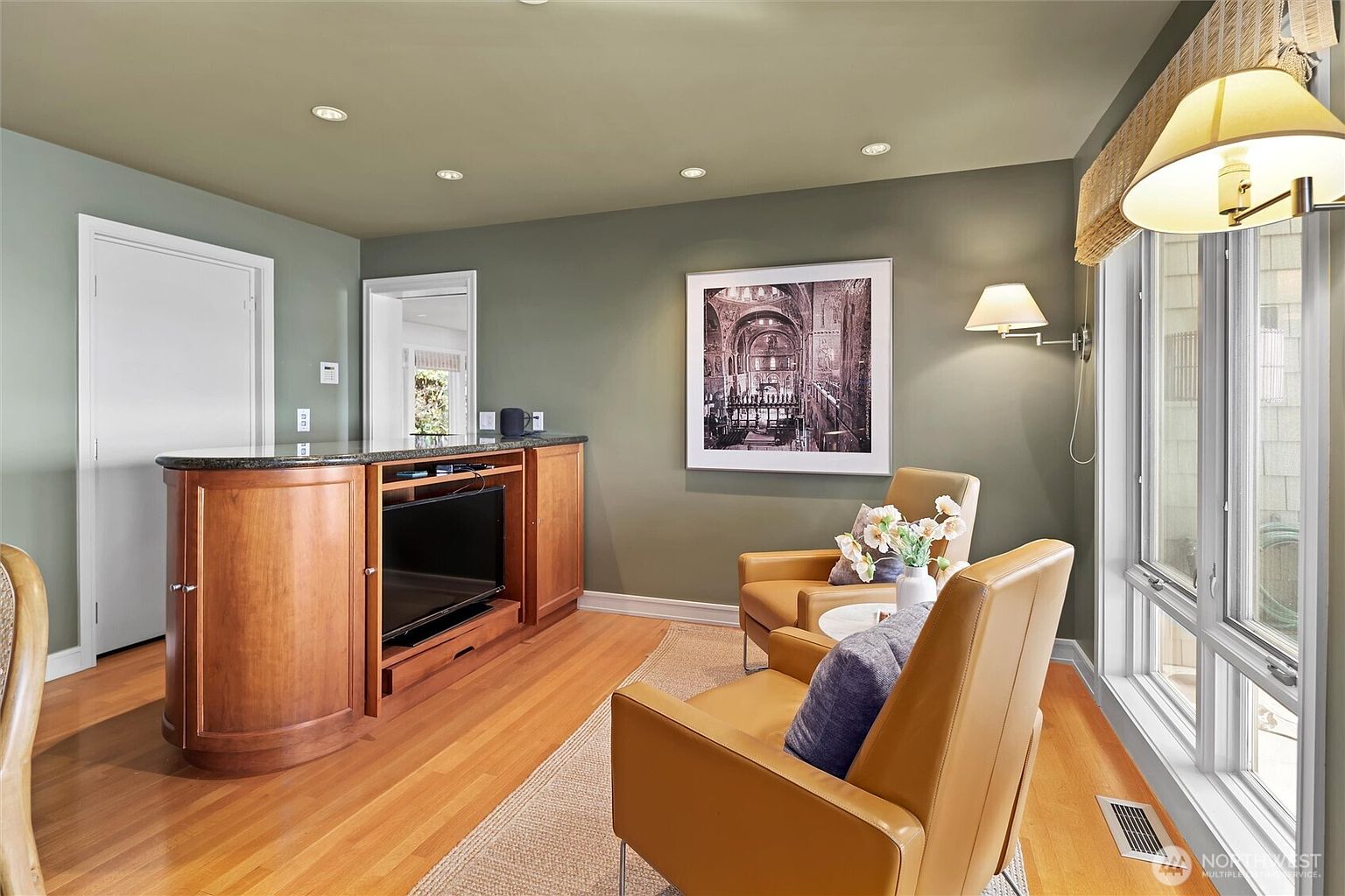 This cozy living area features warm hardwood flooring, sage green walls, and a built-in wooden media console topped with a dark granite counter. Two tan leather armchairs are arranged on a textured area rug, complemented by a wall-mounted sconce and a large framed architectural print. The space is bathed in natural light from a large window, creating an inviting and comfortable atmosphere.