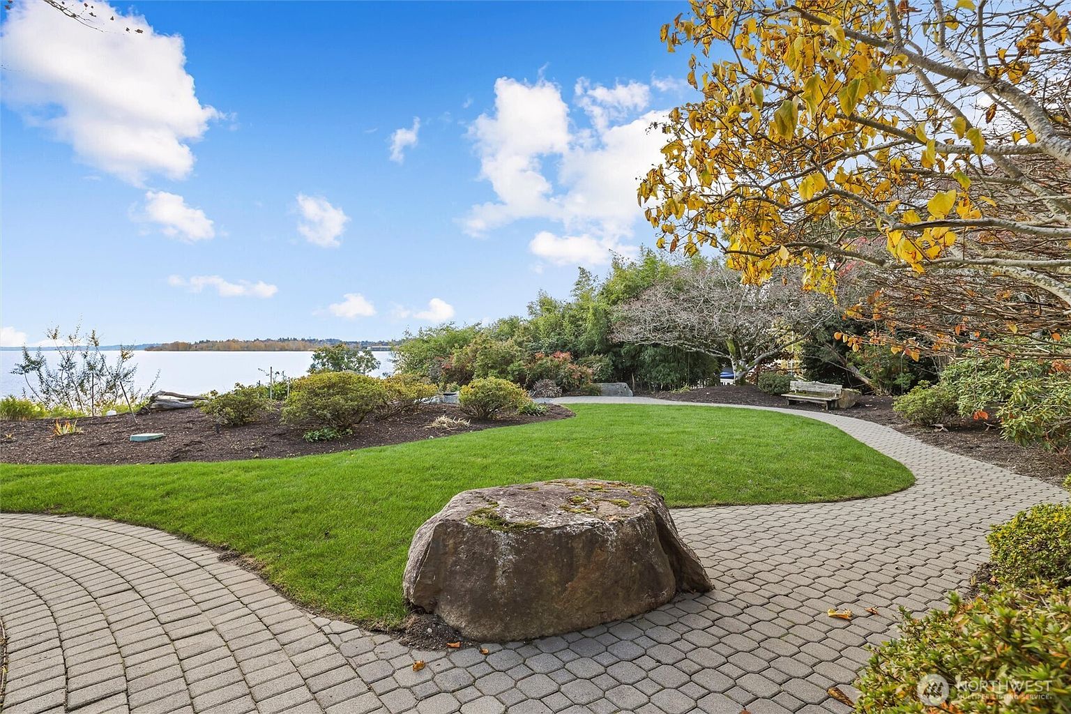 This serene backyard garden features a manicured lawn bordered by a curved paver walkway and a large, natural stone focal point. The landscape includes lush greenery, mature trees with autumn-toned leaves, and a wooden bench, all set against a picturesque waterfront backdrop. The perspective is a wide-angle, eye-level shot that emphasizes the peaceful, park-like atmosphere of the property.