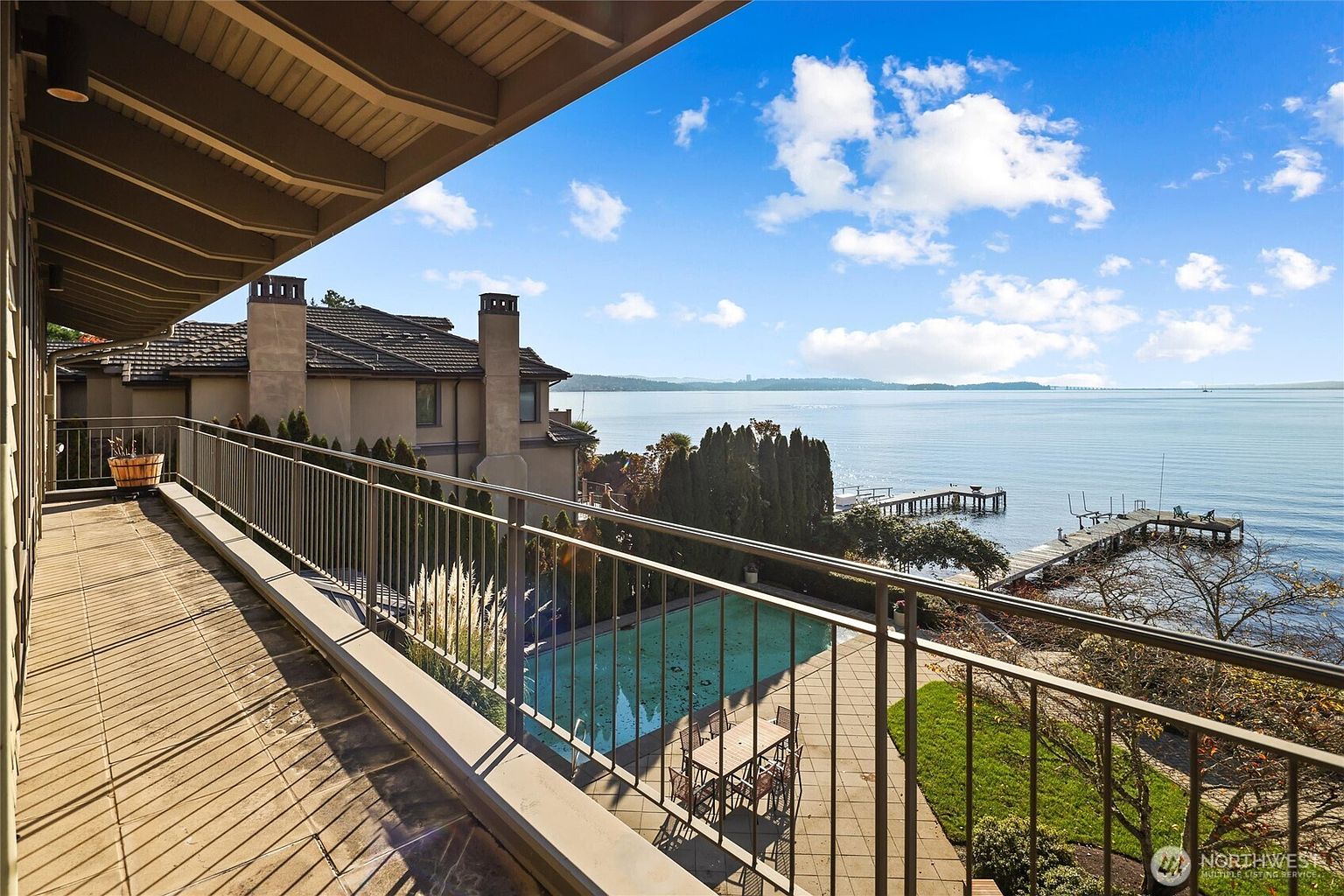 This elevated balcony offers a stunning, panoramic view of a serene lake, complete with a private dock and a sparkling swimming pool below. The wooden deck features a metal railing and provides a perfect vantage point for enjoying the waterfront scenery and the expansive sky. The perspective is from an upper-level balcony, capturing the luxurious outdoor living space and the tranquil natural surroundings.