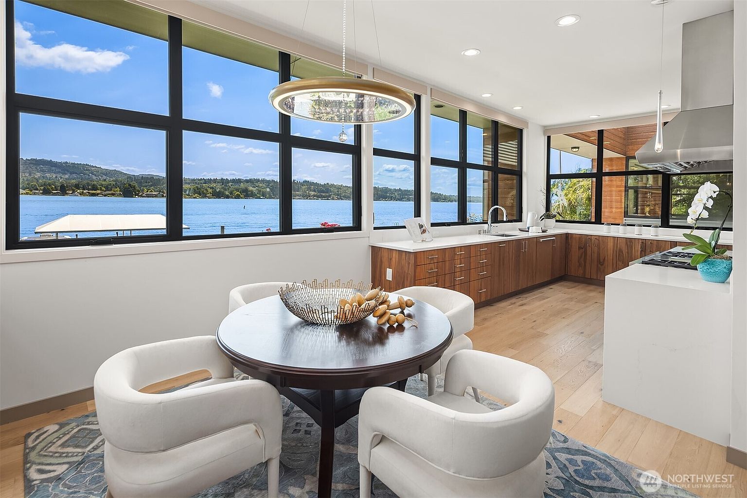 This modern, open-concept kitchen features warm wood cabinetry, sleek white countertops, and a large center island, seamlessly blending into a dining area. Expansive floor-to-ceiling windows provide a stunning, panoramic view of the lake, flooding the space with natural light. The room is anchored by a sophisticated circular dining table and contemporary white chairs, creating an inviting and luxurious atmosphere.