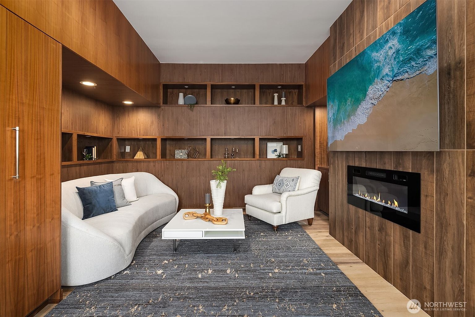 This sophisticated living room features rich, floor-to-ceiling wood paneling that creates a warm and inviting atmosphere. The space is anchored by a modern curved sofa, a white coffee table, and a plush armchair, all set atop a textured dark area rug. A sleek, wall-mounted electric fireplace sits beneath a large, vibrant coastal-themed artwork, while built-in shelving provides elegant display space for decor.