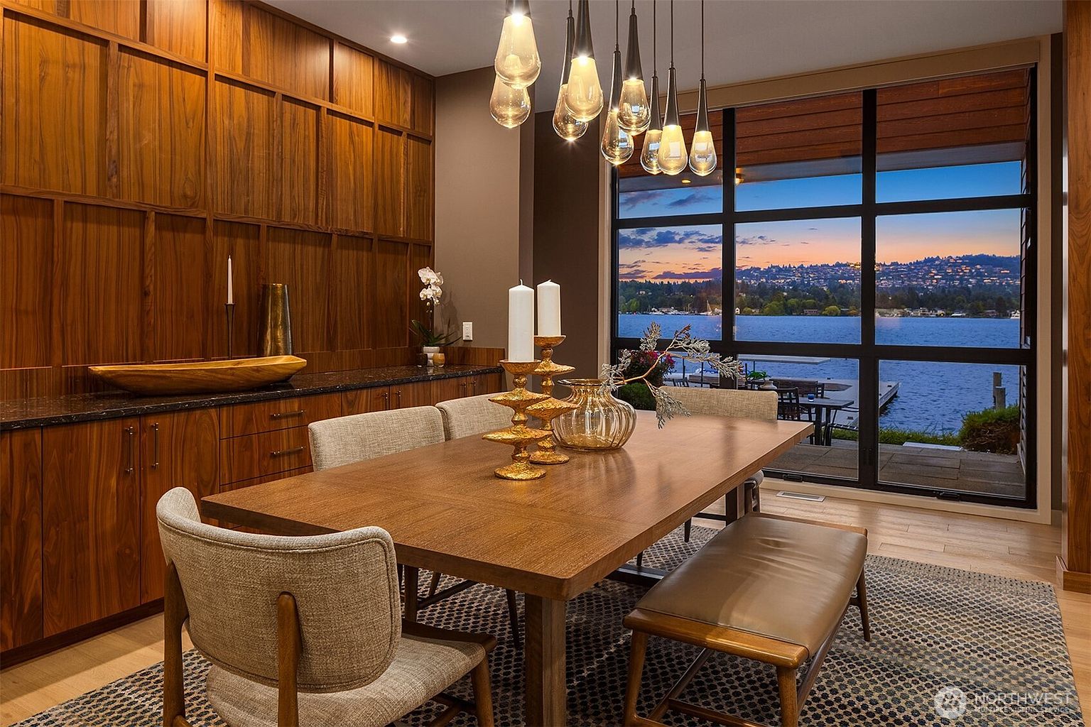 This elegant dining room features a warm, wood-paneled accent wall and a large wooden table paired with upholstered chairs and a leather bench. A striking cluster of teardrop-shaped pendant lights hangs above the table, while floor-to-ceiling windows provide a breathtaking, panoramic view of a lake at sunset. The space exudes a sophisticated, modern aesthetic, perfectly blending natural textures with a serene waterfront backdrop.