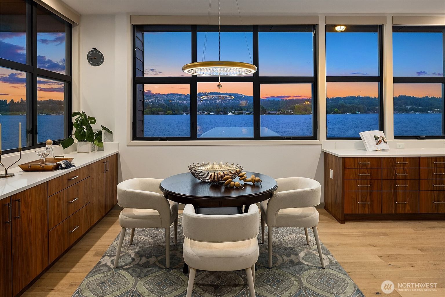 This elegant dining area features a round dark wood table surrounded by four cream-colored upholstered chairs, all set atop a patterned area rug. The room is framed by large windows that offer a breathtaking, panoramic view of a lake at sunset, complemented by a modern circular chandelier. Flanking the dining space are custom wood cabinets with white countertops, creating a sophisticated and functional atmosphere.