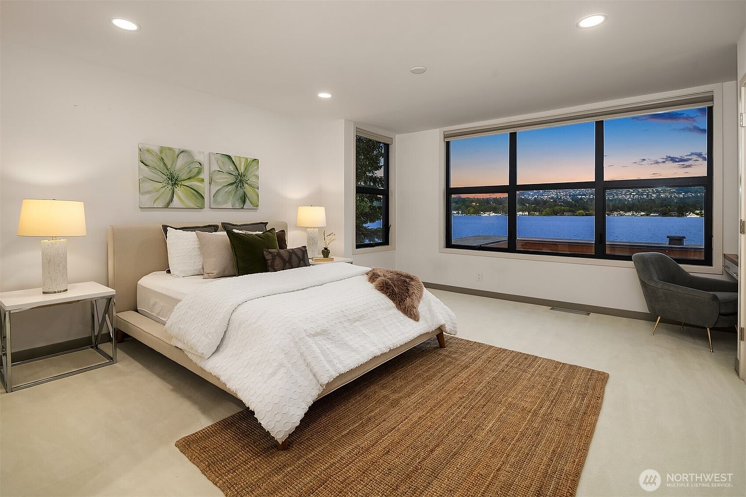 This spacious primary bedroom features a modern, minimalist aesthetic with a neutral color palette and a plush, textured area rug. A large window provides a stunning, panoramic view of the lake at sunset, while a sleek bedside table and a contemporary armchair add functional elegance. The room is well-lit with recessed lighting, creating a serene and inviting atmosphere.