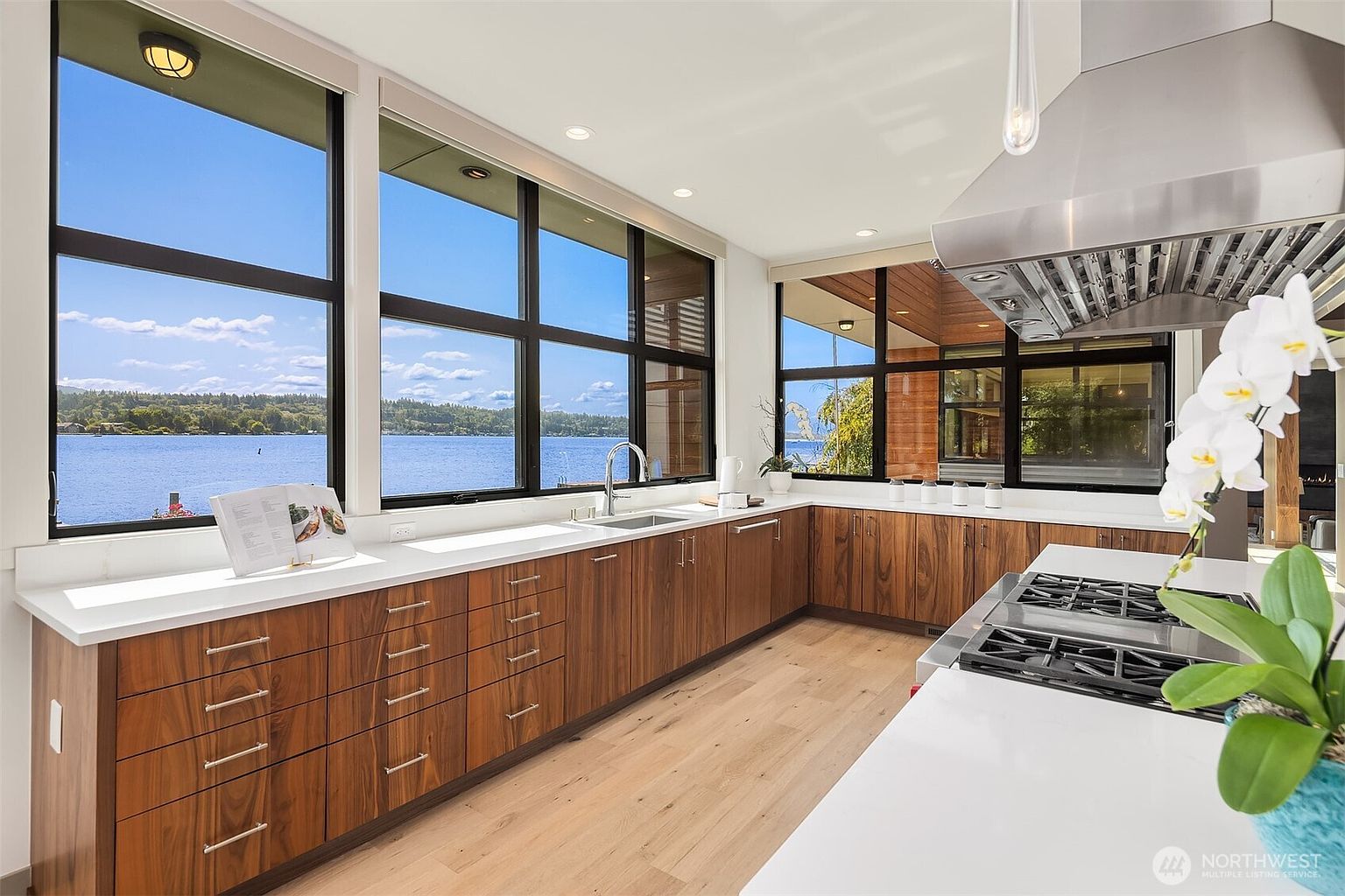 This modern, high-end kitchen features sleek walnut cabinetry paired with crisp white countertops and a large central island. Expansive floor-to-ceiling windows provide a stunning, panoramic view of the lake, flooding the space with natural light. The minimalist design is complemented by professional-grade stainless steel appliances, creating a sophisticated and airy atmosphere perfect for luxury living.