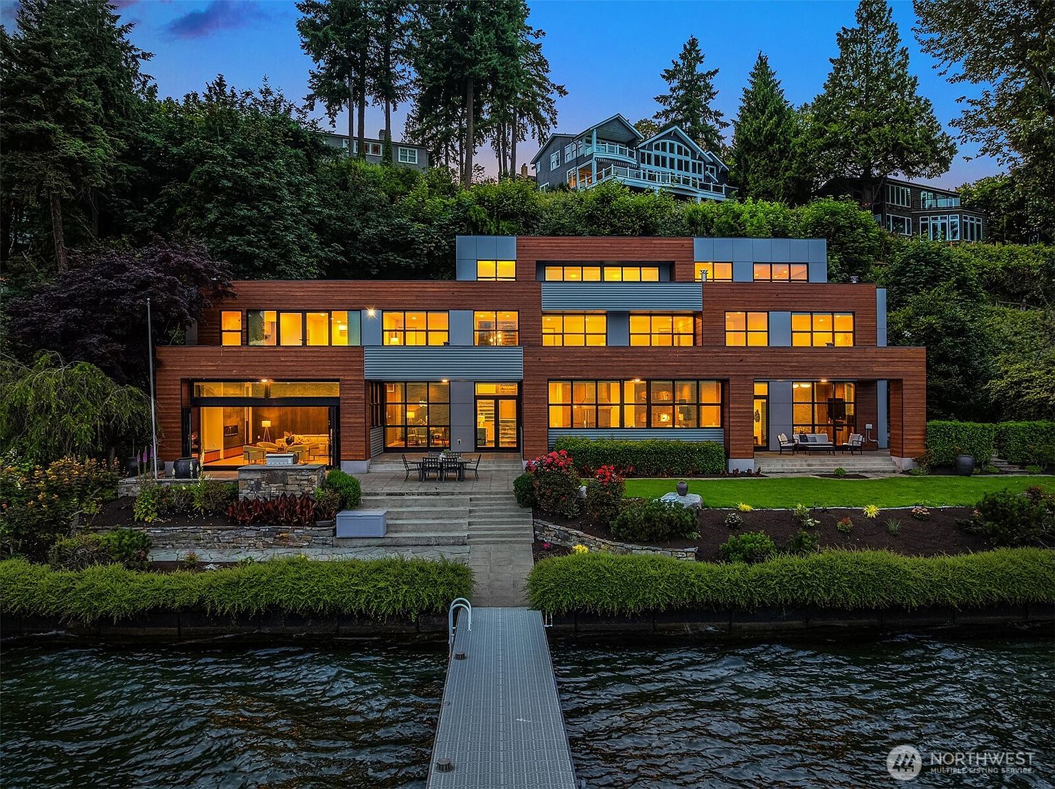 This stunning contemporary waterfront estate features a multi-level design with warm wood siding and expansive floor-to-ceiling windows that capture the evening light. The property includes a private dock extending into the water, a manicured lawn, and a stone patio area perfect for outdoor entertaining. The architectural style is sleek and modern, set against a lush, forested hillside backdrop.