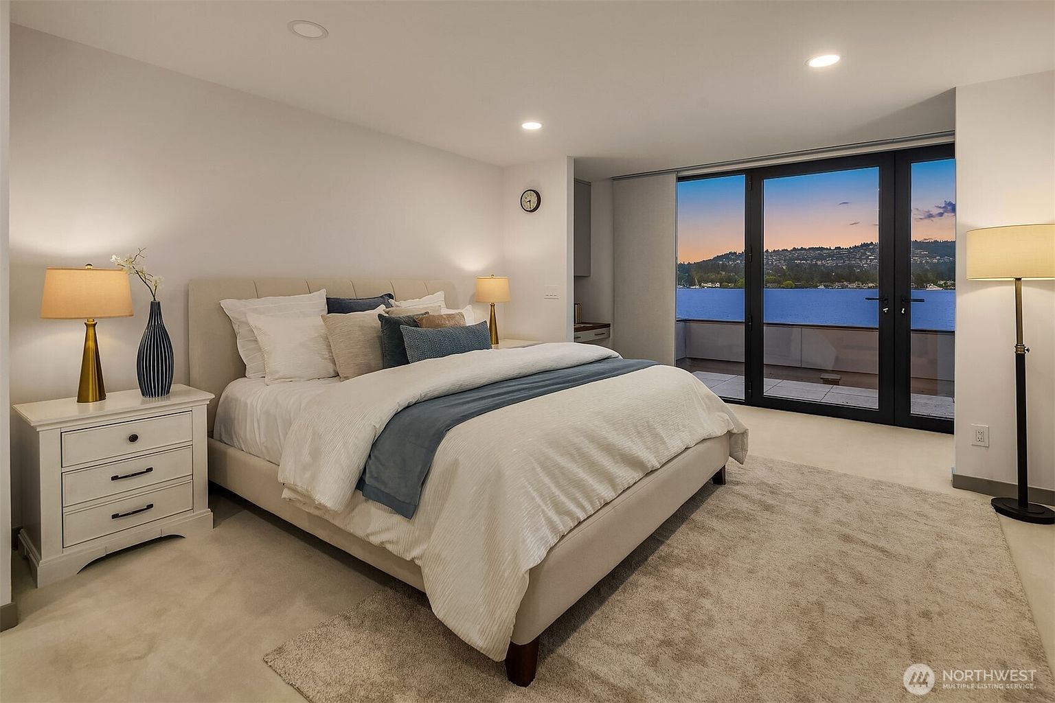 This elegant primary bedroom features a plush, neutral-toned bed as the centerpiece, complemented by a soft area rug and matching bedside lamps. Large floor-to-ceiling glass doors open onto a private balcony, offering a stunning, unobstructed view of the water and sunset. The room exudes a serene and sophisticated atmosphere, perfect for a luxury waterfront residence.