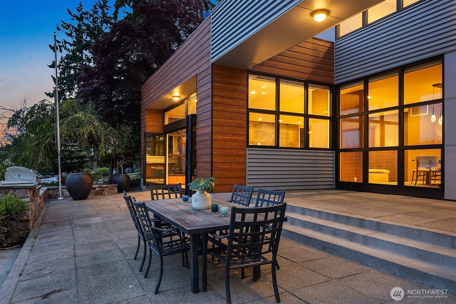 This image showcases a modern, multi-level outdoor patio space featuring a dark metal dining set and a stone-paved floor. The architecture blends warm wood siding with sleek, industrial-style metal panels and expansive floor-to-ceiling windows that glow with warm interior lighting. The perspective is a low-angle shot that emphasizes the seamless transition between the indoor living area and the inviting, sophisticated outdoor entertainment space.