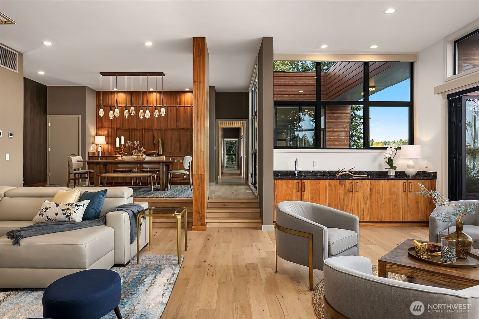 This sophisticated open-concept living space features a sunken dining area with warm wood paneling and a modern chandelier, seamlessly connected to a bright living room with light hardwood floors. A sleek wet bar with dark stone countertops and natural wood cabinetry sits adjacent to large windows that invite abundant natural light. The overall aesthetic is a blend of mid-century modern warmth and contemporary luxury, creating an inviting and airy atmosphere.