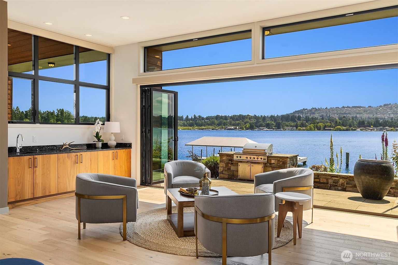 This sophisticated indoor-outdoor living space features a seamless transition to a waterfront patio through large, folding glass doors. The interior is furnished with four modern, grey upholstered armchairs arranged around a central coffee table on a circular rug, complemented by a sleek wet bar with light wood cabinetry and dark countertops. The expansive windows frame a stunning, unobstructed view of the lake and distant hills, creating a serene and luxurious atmosphere.