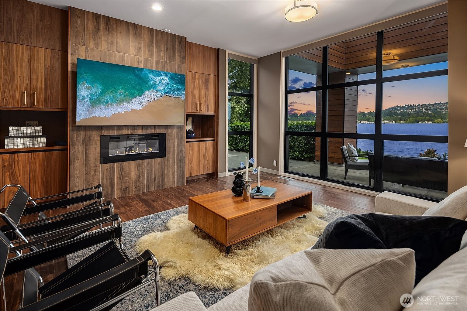 This sophisticated living room features a striking wood-paneled accent wall with a built-in fireplace and a large flat-screen television. Floor-to-ceiling windows provide a panoramic view of the lake, seamlessly blending the modern interior with the natural beauty outside. The space is furnished with mid-century modern chairs, a plush area rug, and a warm wood coffee table, creating an inviting and luxurious atmosphere.