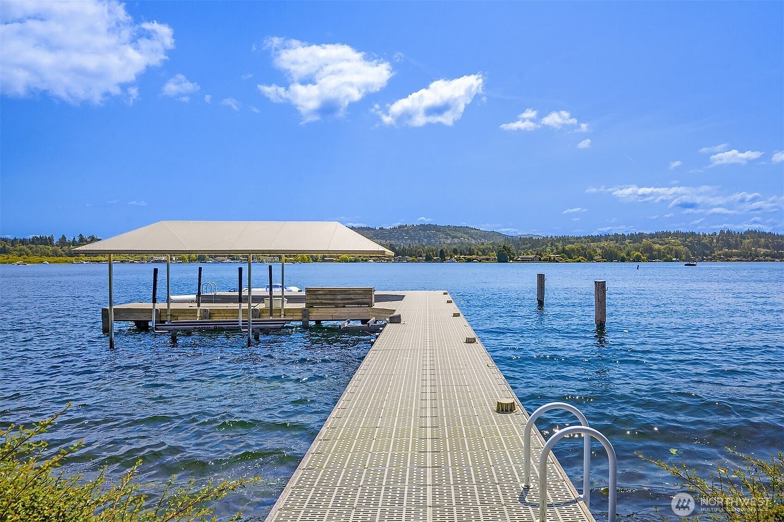 This image captures a serene, elevated perspective of a private dock extending into a calm, expansive lake under a bright blue sky with scattered clouds. The dock features a covered boat slip on the left and a metal ladder on the right, providing direct access to the water. The surrounding landscape includes distant hills and lush greenery, creating a peaceful and inviting waterfront atmosphere perfect for recreational activities.