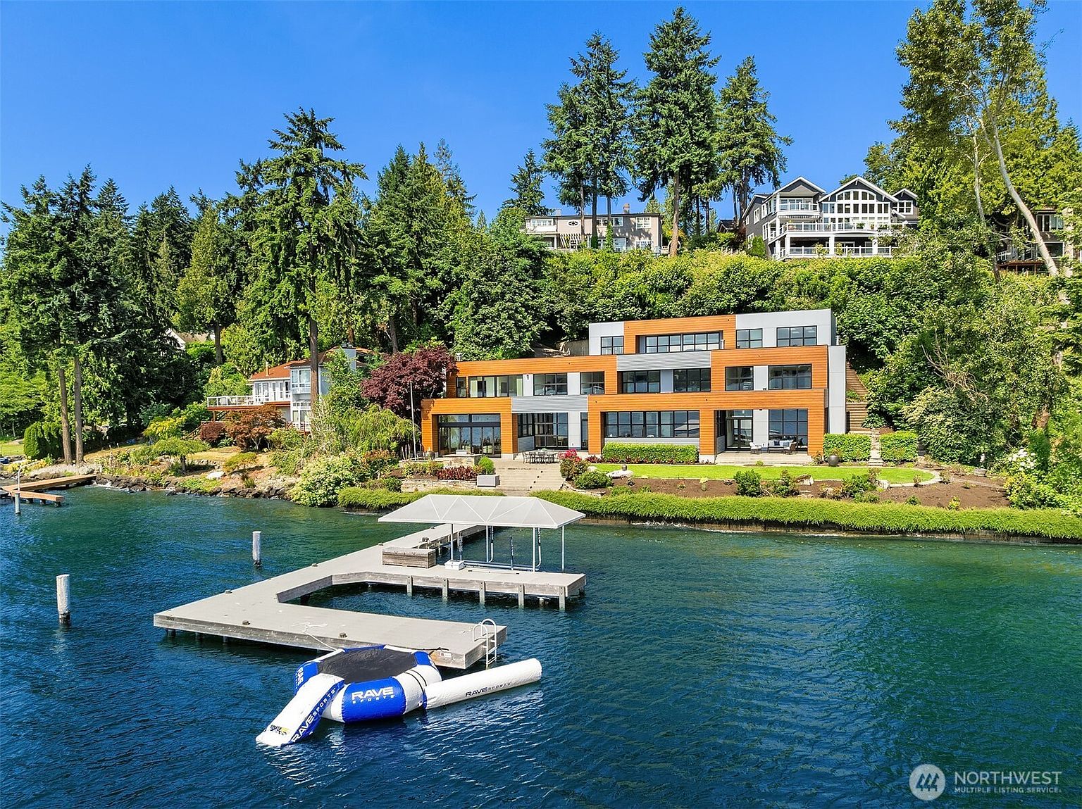 This stunning aerial view captures a modern, multi-level waterfront estate nestled against a lush, tree-covered hillside. The home features a contemporary design with wood-paneled accents and expansive glass windows, complemented by a private dock extending into the deep blue water. The property offers a seamless blend of luxury architecture and natural beauty, creating a serene and exclusive lakeside atmosphere.
