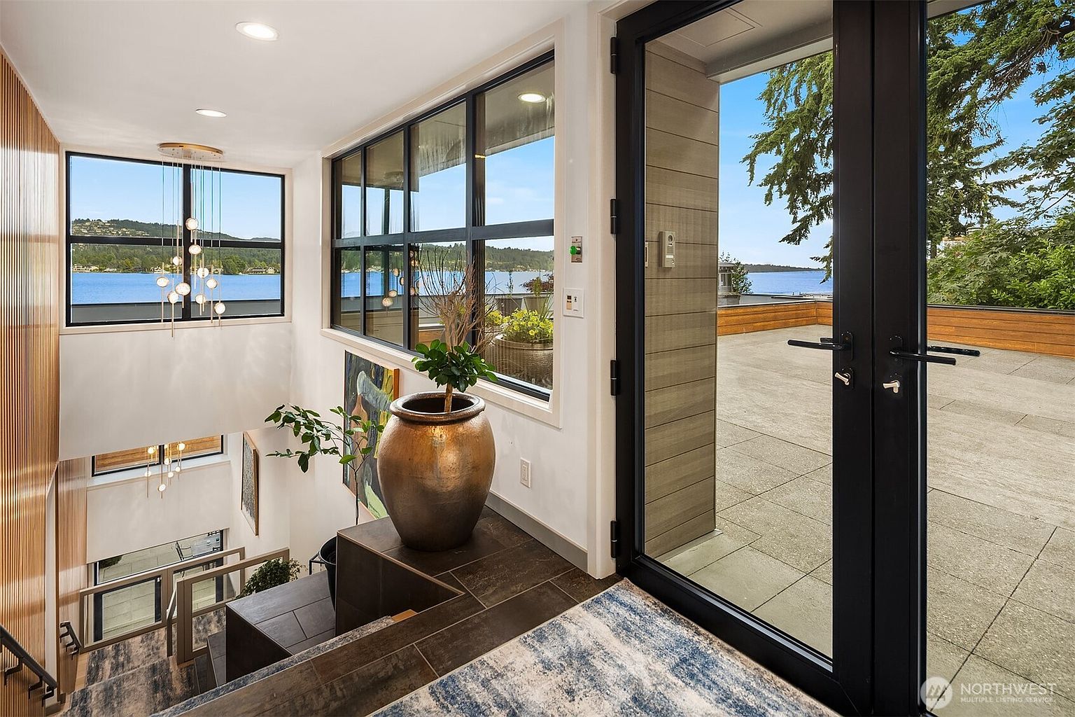 This sophisticated entryway and staircase area features a modern, open-concept design with a striking view of the lake through expansive windows. A large, metallic decorative urn sits on a tiled landing, while a contemporary chandelier hangs above the staircase, creating a sense of luxury and depth. The seamless transition between the interior space and the outdoor deck through black-framed glass doors emphasizes the home's connection to its scenic surroundings.