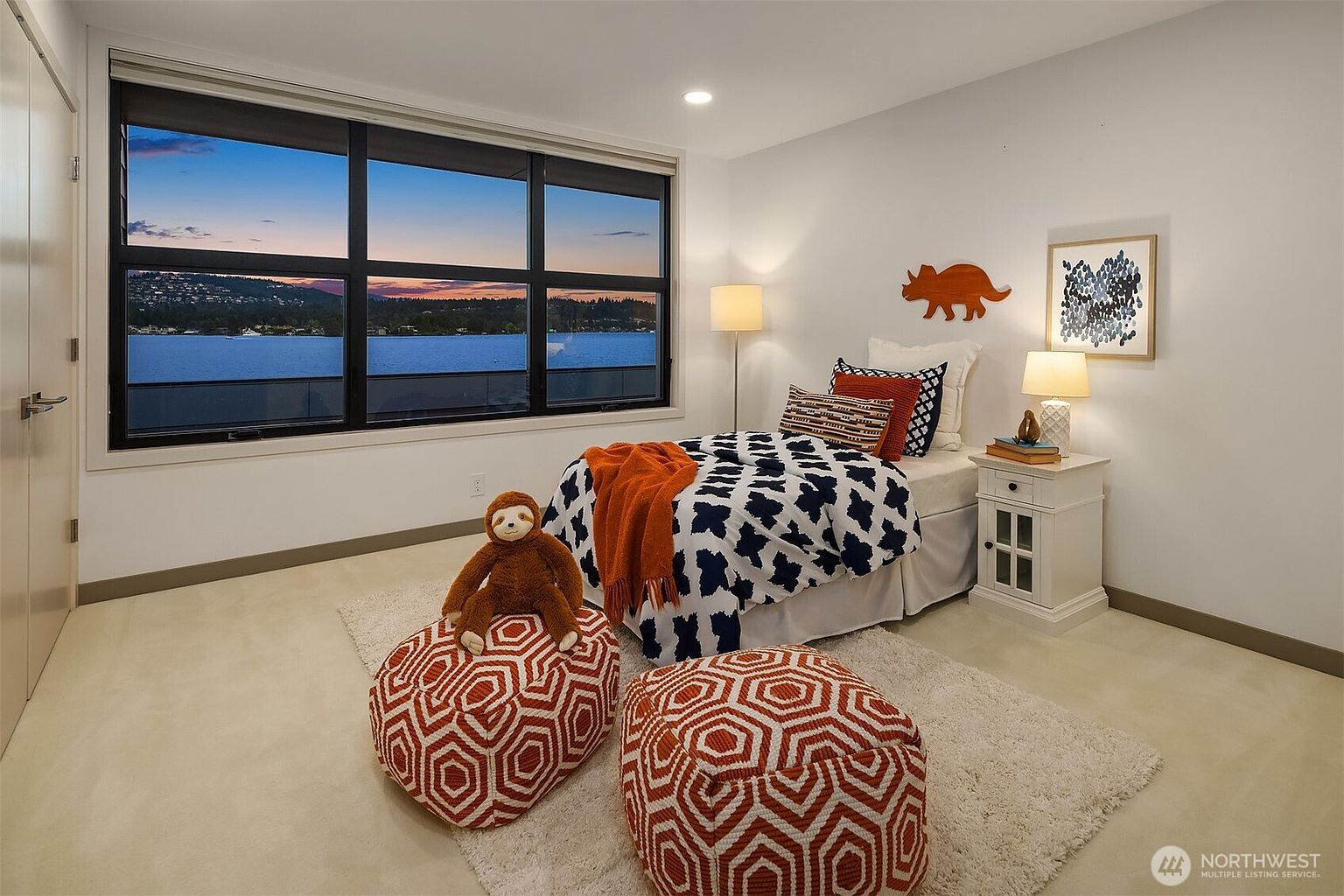 This cozy, well-lit bedroom features a twin bed with a bold navy and white patterned comforter, accented by a vibrant orange throw blanket and matching geometric poufs. A large window provides a stunning, panoramic view of the lake at sunset, while a playful dinosaur wall decoration and a stuffed sloth add a whimsical touch. The room is finished with neutral carpeting and a classic white nightstand, creating a warm and inviting atmosphere.
