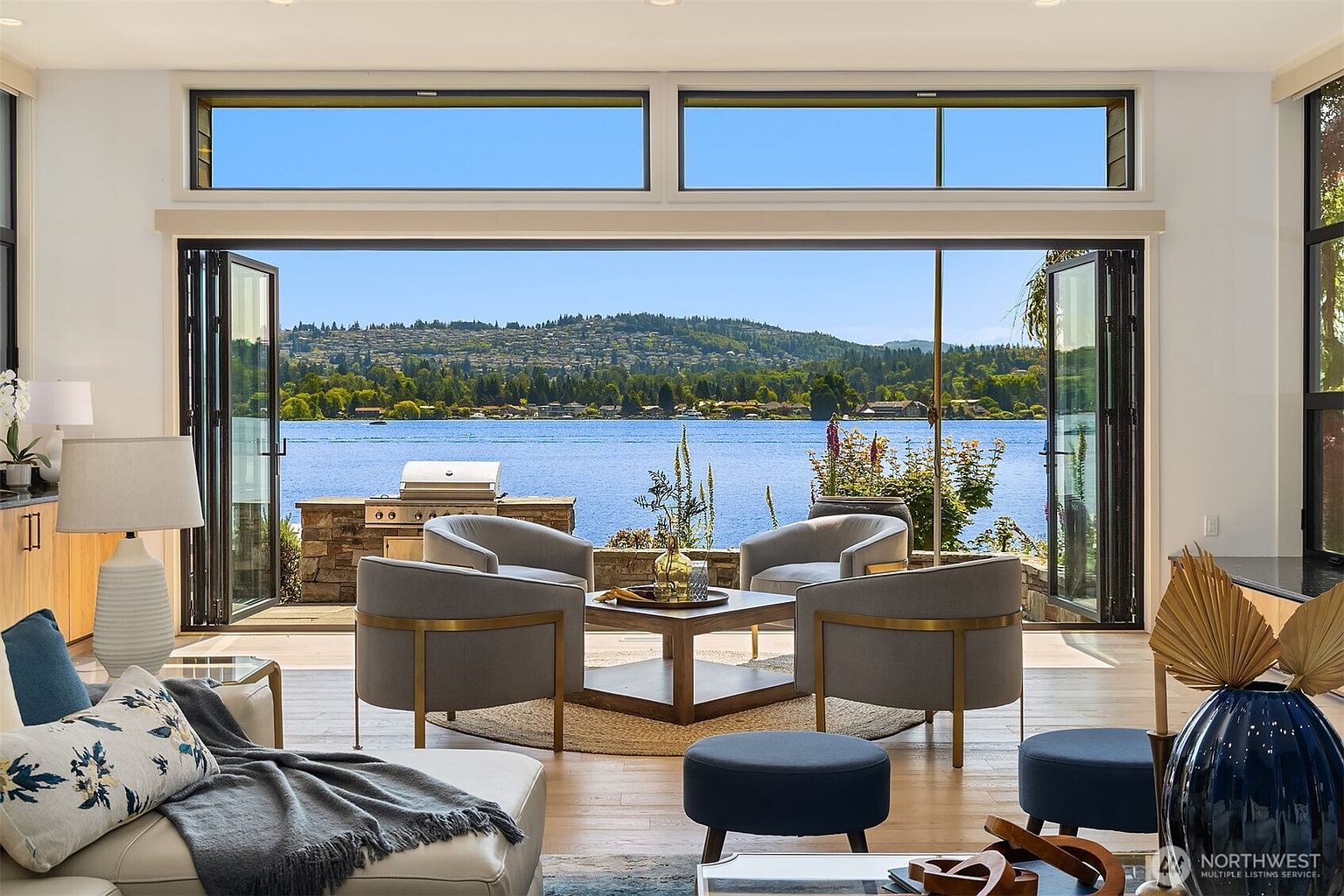 This sophisticated living room features an open-concept design that seamlessly transitions to an outdoor patio through expansive, floor-to-ceiling folding glass doors. The space is furnished with elegant grey armchairs arranged around a central wooden coffee table, all set against a stunning, unobstructed view of a serene lake and distant hills. The interior aesthetic is modern and airy, emphasizing a strong connection between indoor comfort and the natural beauty of the waterfront setting.