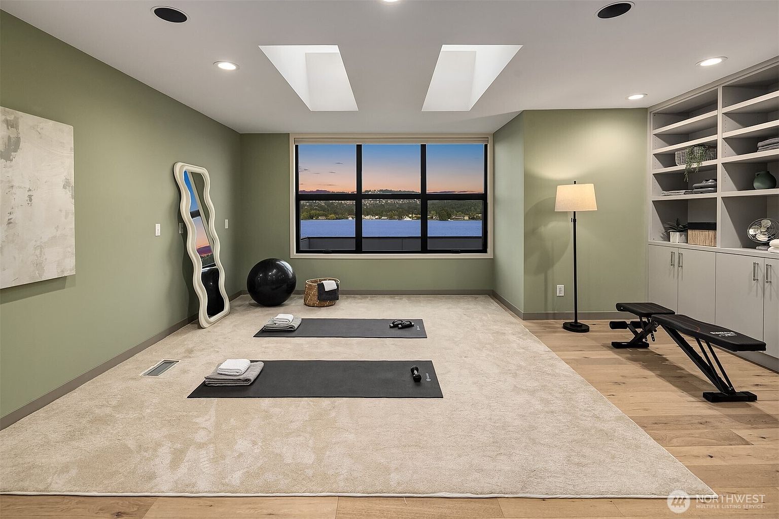 This bright and airy home gym features sage green walls, light hardwood flooring, and a large window offering a serene view of the water at sunset. The space is equipped with two yoga mats, a weight bench, a full-length wavy mirror, and built-in shelving, creating a tranquil and functional workout environment. The room is illuminated by recessed lighting and two skylights, enhancing the calm and inviting atmosphere.